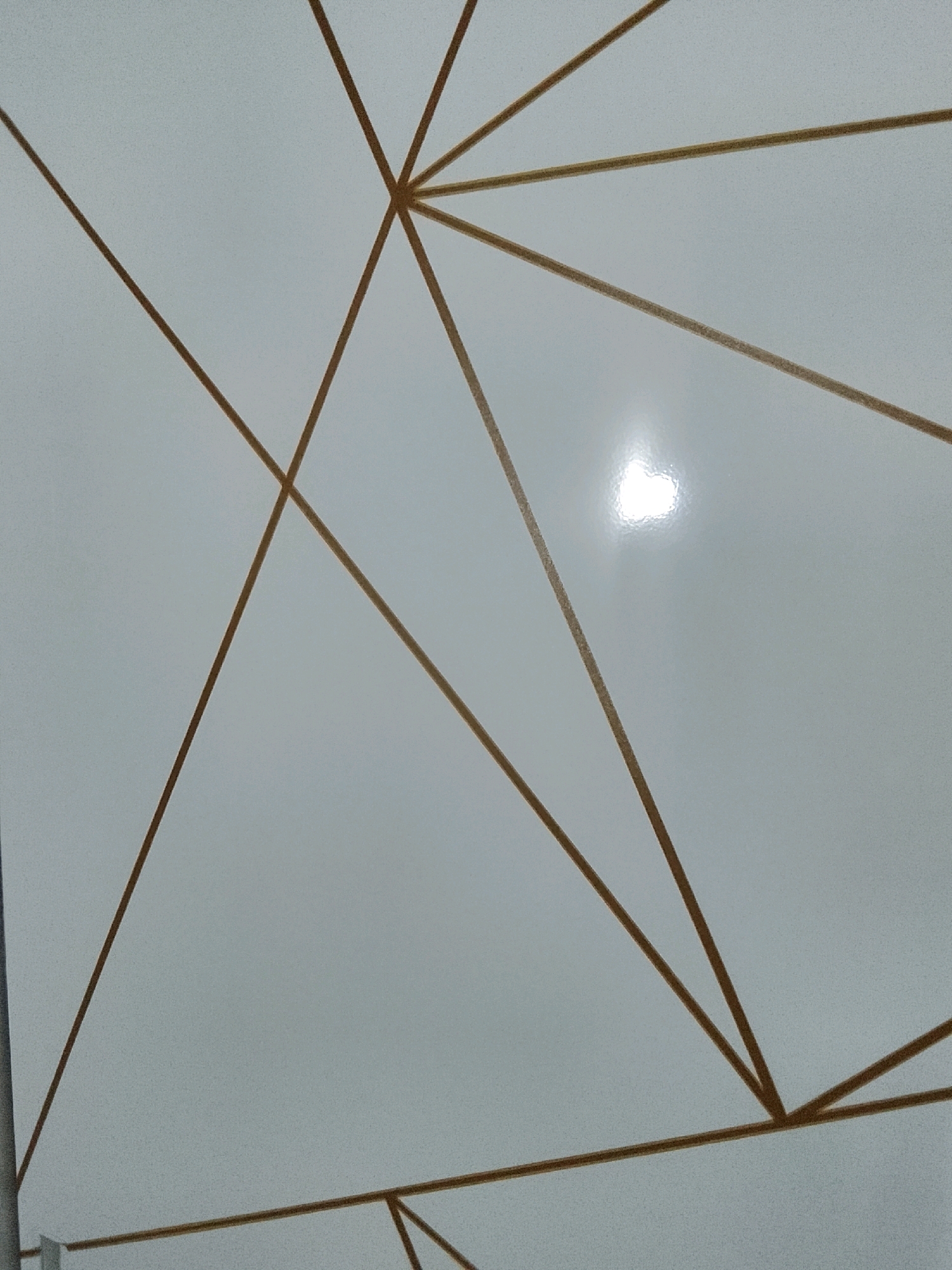 The image displays a white, textured surface with a geometric pattern of thin, intersecting brown lines. The lines form various angles and appear to be part of a larger design. There is a bright, specular reflection visible on the surface, indicating it is smooth and possibly glossy, which could be from a light source. The background is a light, neutral color with subtle graininess. The image does not contain any people, animals, or identifiable objects. It is an abstract pattern on a surface. There is no information in the image to suggest a specific location in Kalibeji, Indonesia.