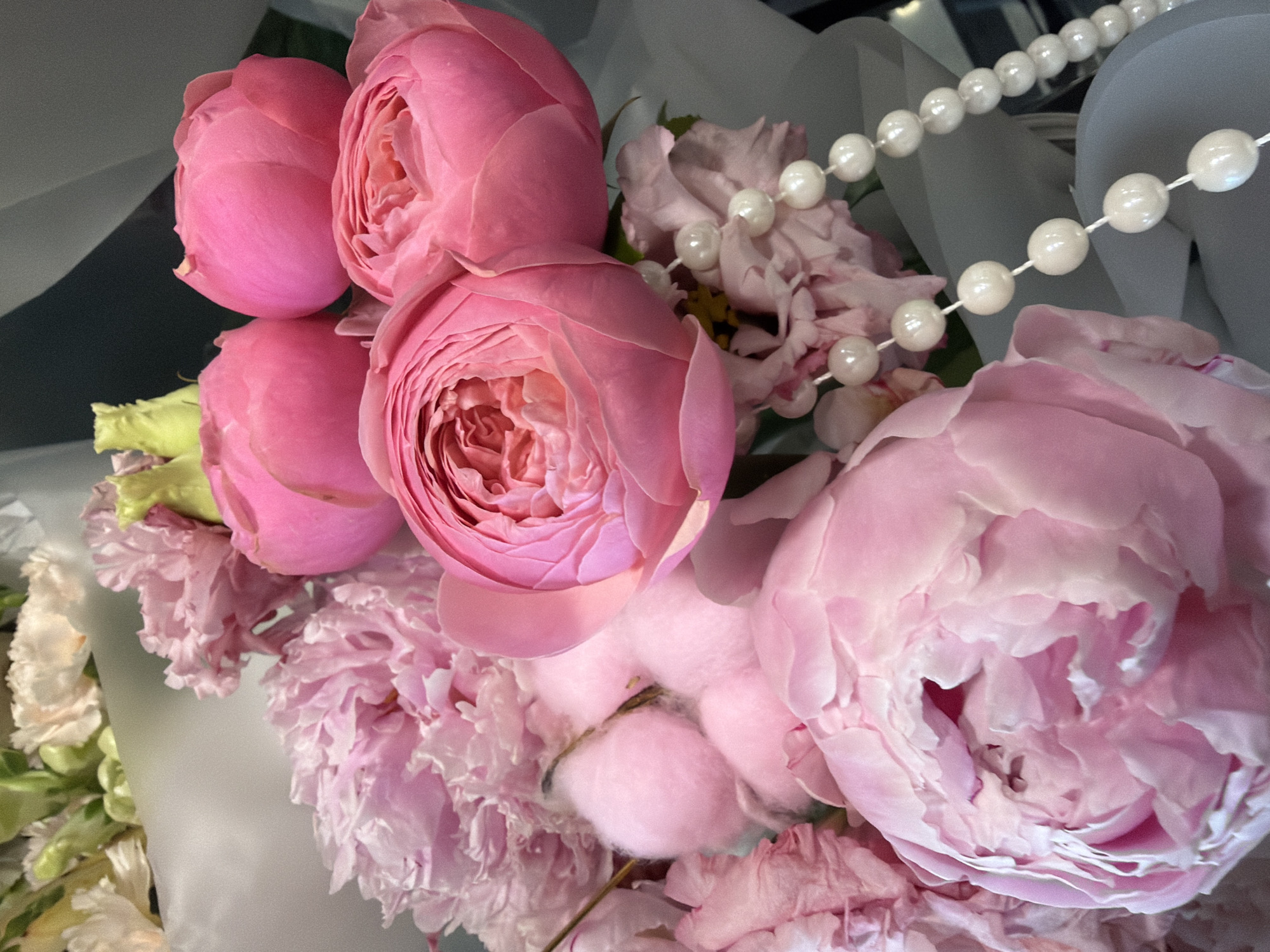 A bouquet of pink flowers is displayed against a dark background. The bouquet features several roses with tightly packed petals, as well as larger, softer peonies. Interspersed among the flowers are cotton balls and a string of white pearls. The lighting suggests an indoor setting, possibly a flower shop or a private residence. The image is a close-up shot, focusing on the textures and colors of the floral arrangement.