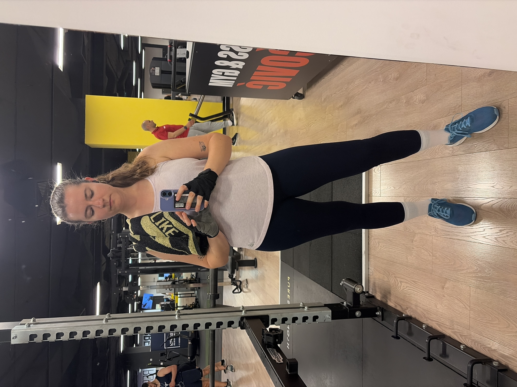 An image, rotated clockwise by 90 degrees, captures a woman taking a mirror selfie inside a gym. The woman, with light skin and brown hair pulled back, is reflected from her chest to her feet. She wears a light-colored tank top, dark leggings, white socks, and blue athletic shoes. She holds a purple smartphone in her left hand, which is covered by a black workout glove. A black and yellow towel, bearing the text "NIKE", is also clutched in her hand.
The background, visible through the mirror's reflection, reveals a gym interior. To the right, a black metal squat rack or bench structure is partially visible. The flooring transitions from a dark, possibly rubberized mat to a lighter wooden surface. In the mid-ground, a large sign with red and white lettering is reflected, displaying "SONIC! GYM & GAIN!" (appearing reversed due to the mirror). Two other individuals are reflected in the background: one man in a red polo shirt and grey pants, and another man partially visible in a dark tank top. The setting is well-lit, typical of an indoor fitness facility.