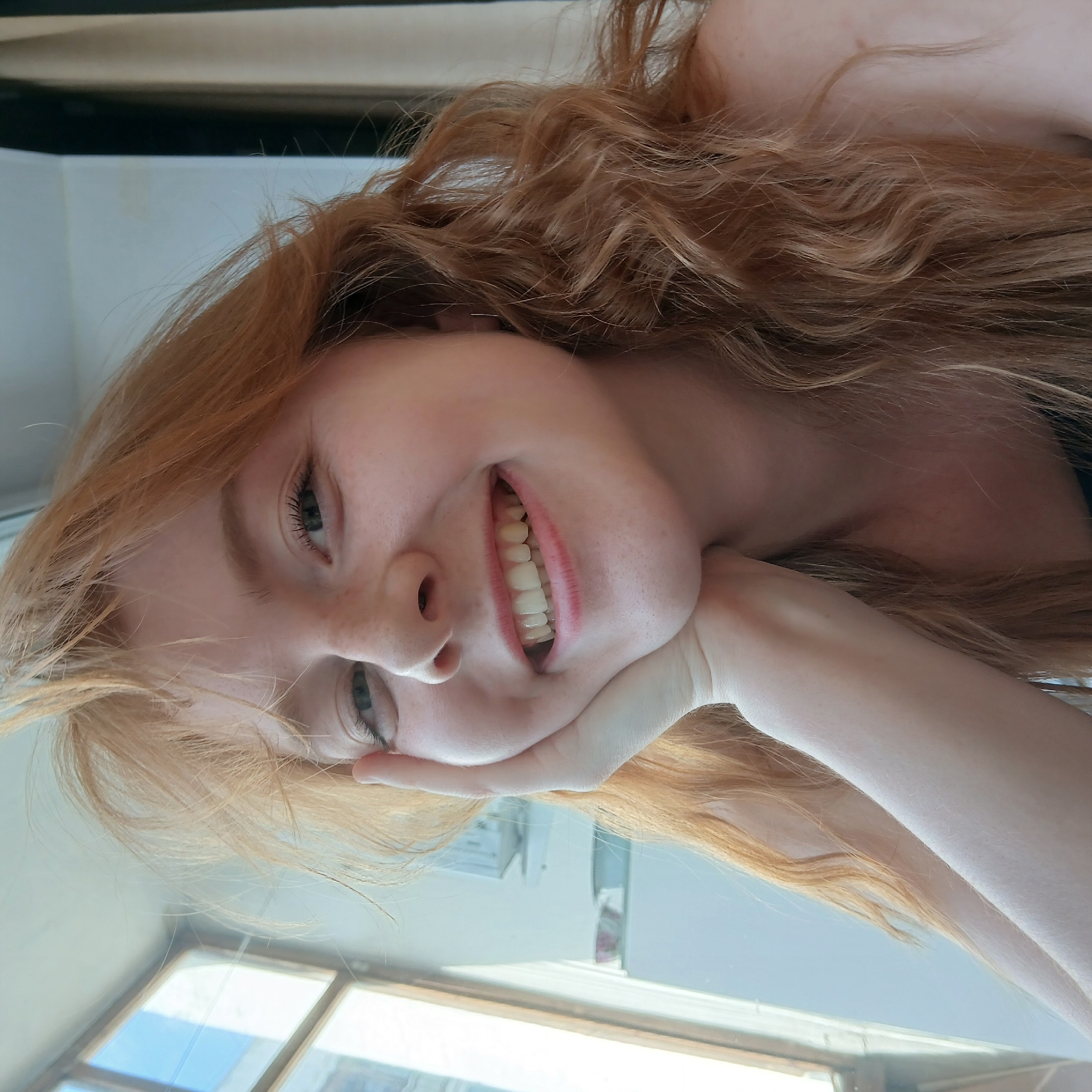 The media file consists of a main image and several close-up crops of it. The main image features a young woman with long, wavy, reddish-blonde hair. She has fair skin with visible freckles on her nose and cheeks, and light-colored eyes. She is smiling broadly at the camera, revealing her teeth, and her left hand is resting under her chin, supporting her face. A dark strap, possibly from clothing, is visible on her right shoulder.
The image is composed at an angle, with the woman's head positioned on the left side of the frame and her body extending towards the right. This angle, combined with the blurred background, suggests it might be a selfie taken from a reflective surface, like a mirror, or simply an intentionally tilted shot.
The woman's expression conveys happiness and contentment, with her wide, genuine smile.
The background, visible as a reflection or through a window, suggests an indoor setting during the daytime, characterized by bright light. Blurred elements in the background include what appear to be windows with clear blue sky visible through them, as well as indistinct interior walls and possibly furniture or window frames.
The close-up crops provide further detail:
* The texture and color of her wavy, reddish-blonde hair are clearly visible, showing individual strands and subtle variations in shade.
* Her light eyes, dark eyelashes, and light eyebrows are highlighted.
* Her wide smile with well-aligned teeth and naturally pink lips are shown in detail.
* The fair skin of her hand and arm is visible, along with a small mole or blemish on her neck/shoulder.
* The background crops confirm the presence of bright, daylight-illuminated windows with a view of the sky and internal structures.
There is no visible text in any of the images. The location context provided is Yekaterinburg, Russia.