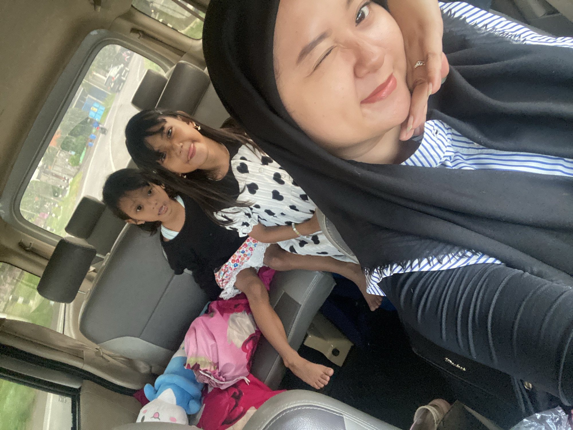 This image captures a selfie taken inside a vehicle, likely a car or van, featuring three individuals: a woman and two girls.
In the foreground, a woman is taking the photo, looking directly at the camera with a playful wink and a slight smile. She is wearing a black hijab, a blue and white striped shirt, and black pants. Her left hand is gently touching her face, revealing a ring on her finger.
Behind the woman, two girls are seated in the vehicle. The older girl, positioned slightly to the left, is smiling and looking towards the camera. She is wearing a black and white top with heart patterns and a colorful patterned skirt. Her bare feet are visible, resting on the seat or items in the car. The younger girl is seated further back, wearing a black top, and appears to be looking forward, not directly at the camera.
The interior of the vehicle shows gray seats and headrests. Through the window on the left side, a road is visible with some distant buildings, trees, and what appear to be street signs, suggesting a daytime setting in an urban or suburban environment. The light indicates it is daytime. On the seat or floor near the girls, there's a blue and white stuffed animal along with various colorful fabrics or blankets. The overall scene conveys a casual and possibly joyful moment of travel, likely in Tlasih, Indonesia. There is no discernible text in the image.