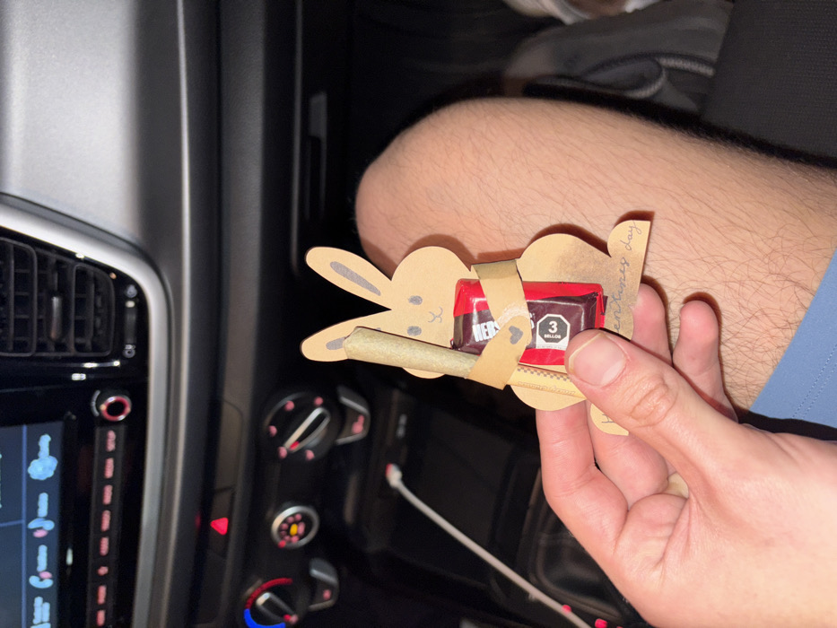 Inside a car, a person holds a crafted paper bunny "basket" containing a Hershey's chocolate mini and a rolled cannabis cigarette. The bunny is light brown with a simple, smiling face. The chocolate is a three-piece mini Hershey's bar. The cannabis cigarette is wrapped in light brown paper. The vehicle's dashboard features climate control knobs and a digital display. Part of a leg and the car seat are visible in the background.