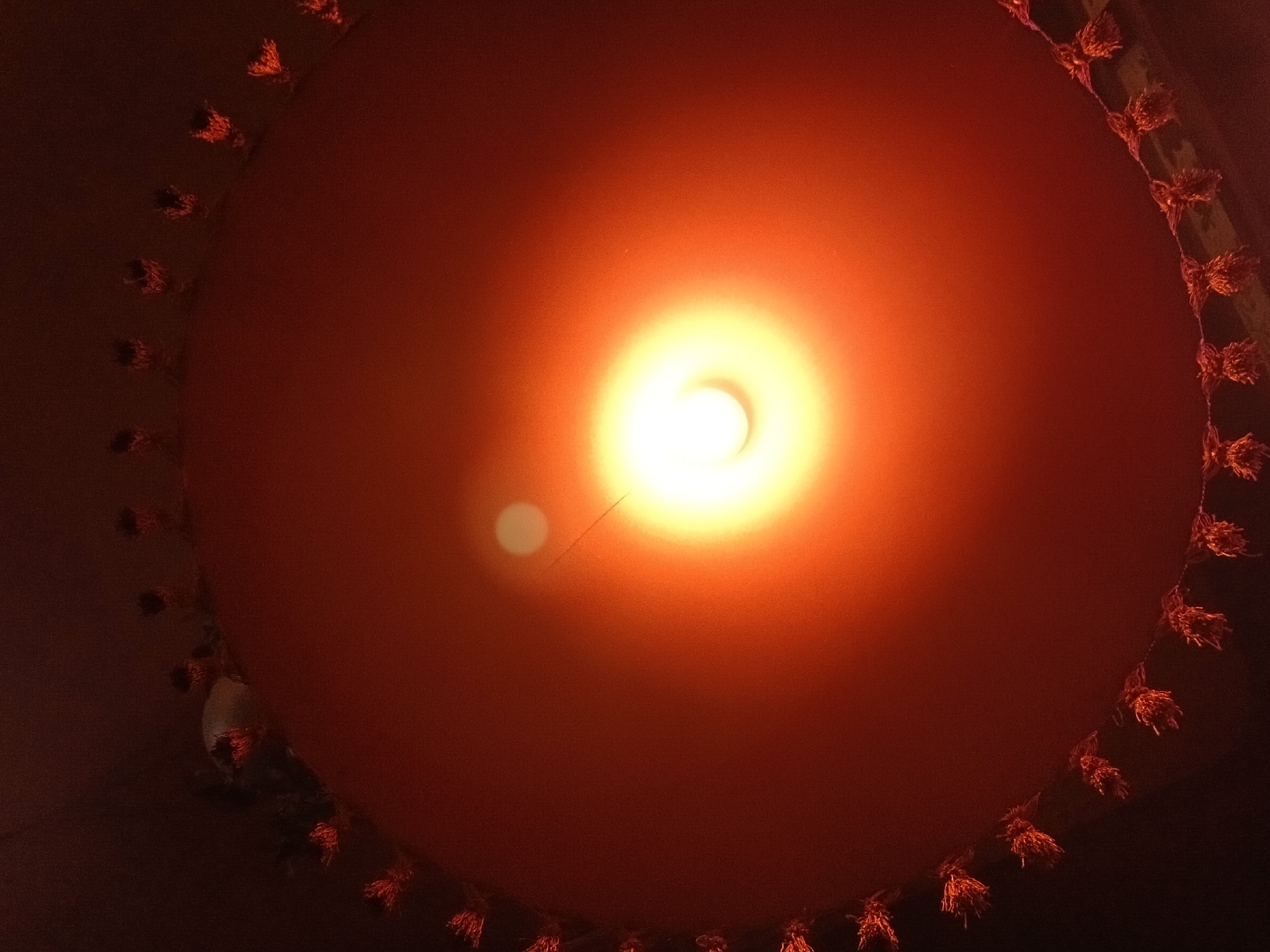 The image is a low-angle shot of a lampshade with a lightbulb illuminated within. The lampshade is orange and has a decorative fringe of small, tassel-like ornaments around its edge. The lightbulb casts a bright glow, creating a halo effect. The background is dark and indistinct. There is no discernible location in the city of Nizhny Novgorod, Russia.