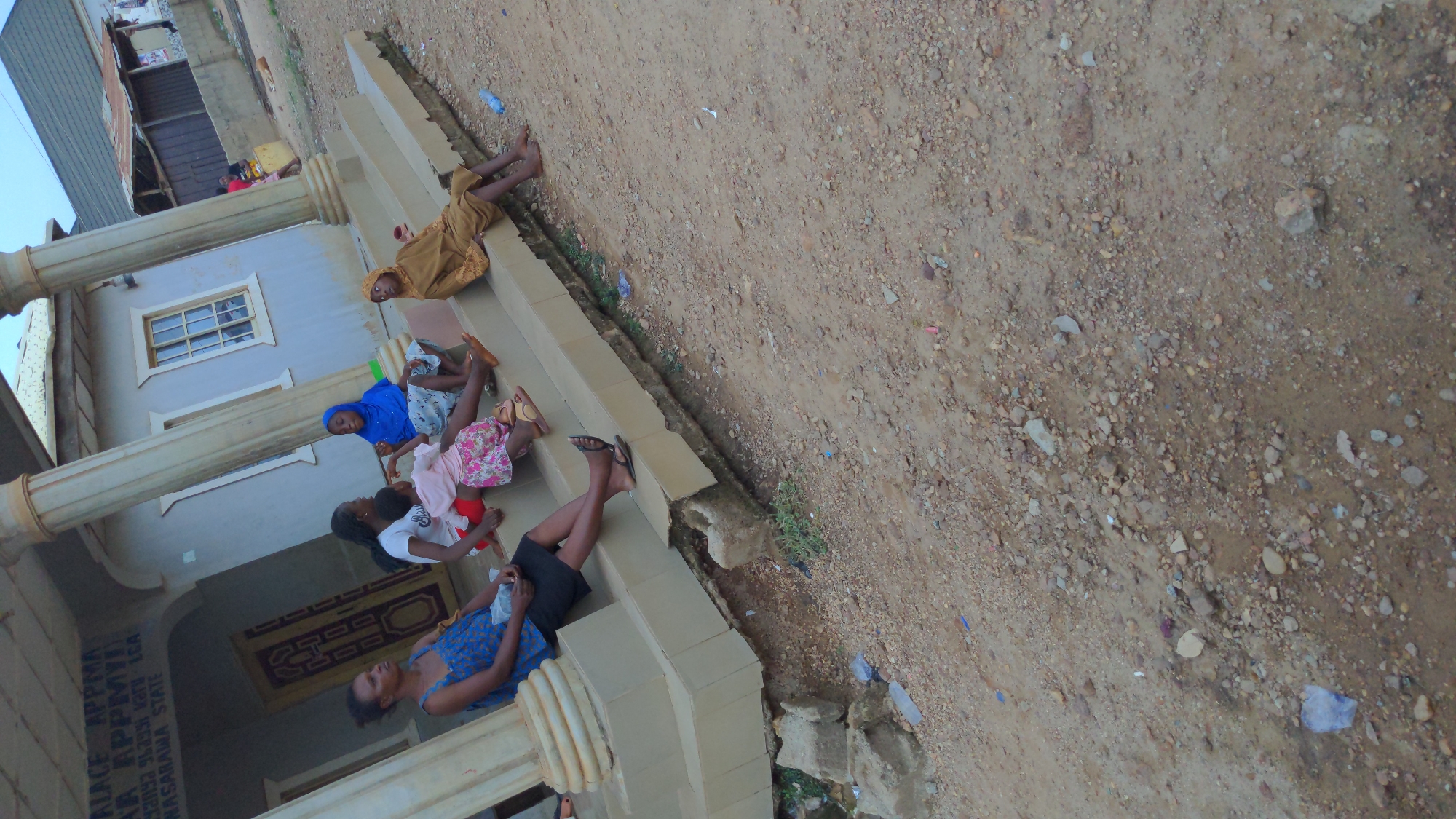 Five people are sitting on the steps of a building. The building has columns and a window with panes. There is dirt and debris on the ground in front of the building. The location is Masaka, Nigeria.