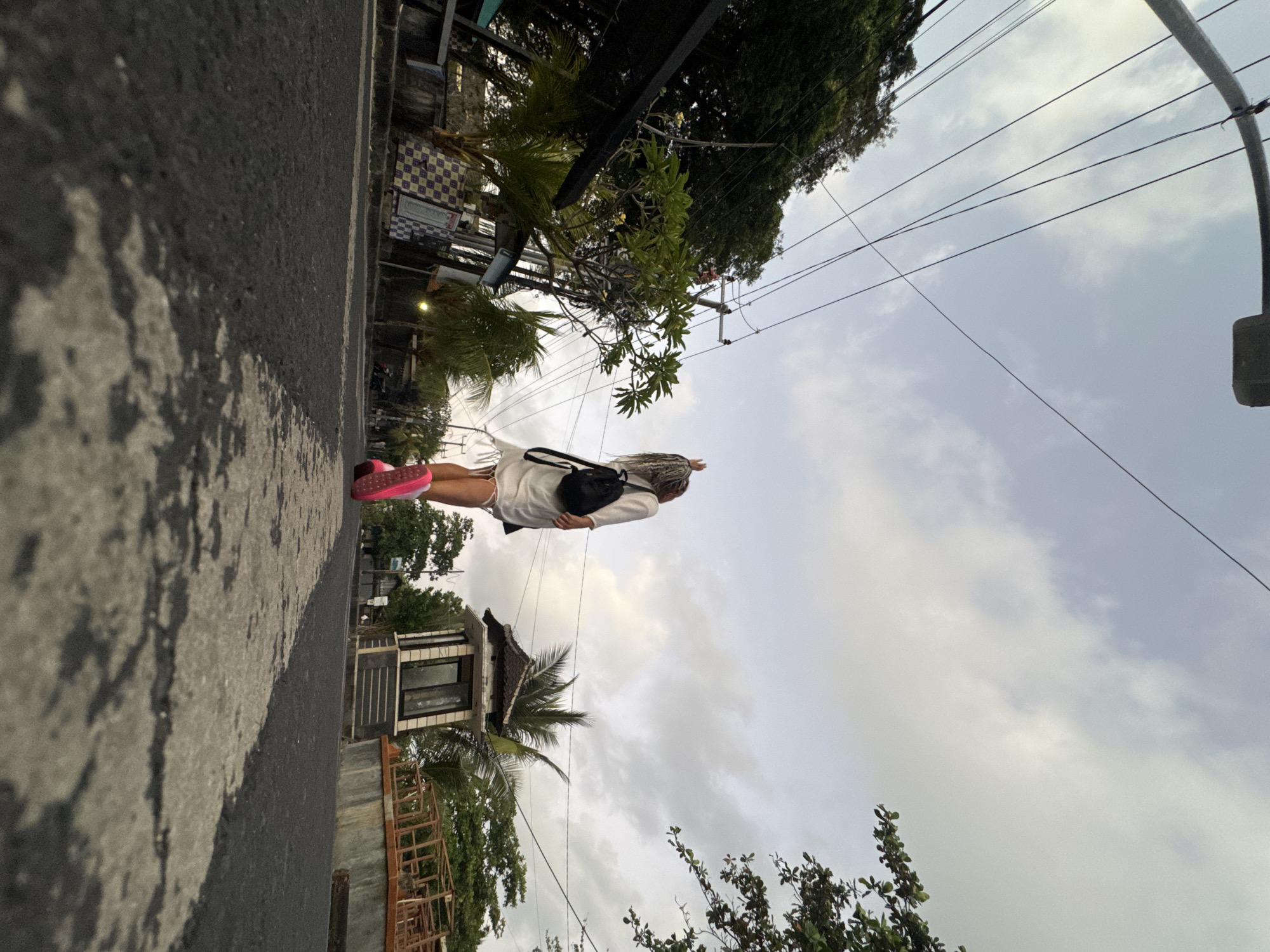 A low-angle shot depicts a person from behind, walking away from the camera on an asphalt road with a white dividing line. The person has long, braided blonde hair and is wearing a white dress, a black backpack, and bright pink shoes. Their right arm is raised, with the hand in a peace sign gesture. Overhead, a network of electrical wires crisscrosses the cloudy sky, supported by utility poles. To the left of the road, lush green trees and some palm trees are visible, along with buildings that appear to be shops or residences. To the right, a small, ornate structure resembling a guardhouse or information booth stands next to a concrete wall. The overall setting suggests a street in Kuta, Indonesia, likely during daytime with overcast weather.