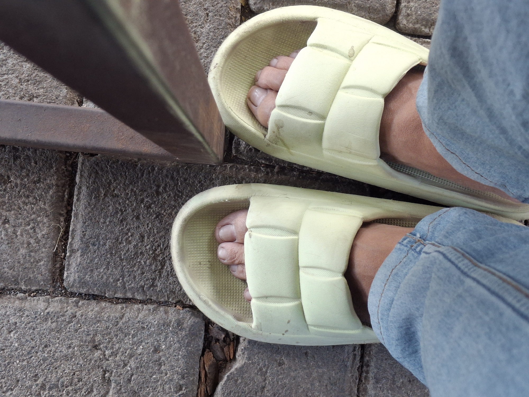 The image shows a first-person perspective of a person's feet wearing light yellow slides, standing on a cobblestone path. The person is wearing light blue jeans. A rusty metal railing is visible to the right, casting a shadow. The scene appears to be outdoors. The cobblestones are of varying sizes and textures, with some dirt and debris in the cracks between them. The slides have a textured interior. The feet are bare within the slides, and the toenails are visible. The lighting suggests it is daytime. This scene is consistent with a common urban or park environment in Lumajang, Indonesia, such as a sidewalk or pathway.