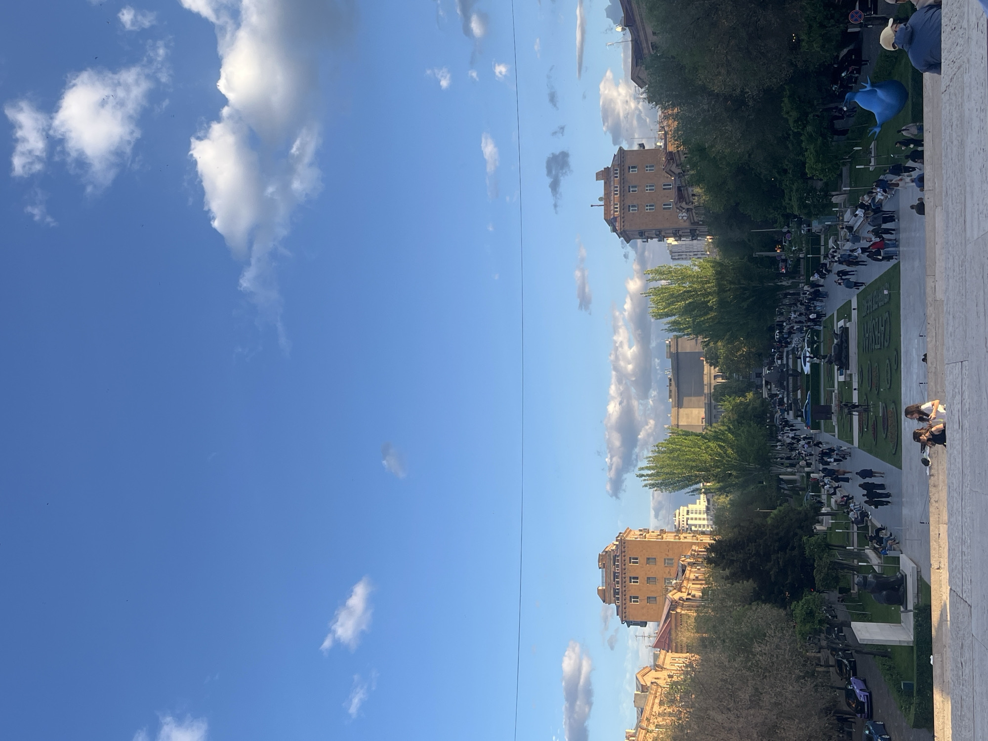 The image shows the Cascade complex in Yerevan, Armenia, viewed from above. A large crowd of people is visible, walking along the landscaped promenade. Several buildings are seen alongside the promenade, and trees line the street. A sculpture of a blue bird is prominent. The sky is blue with scattered clouds. An overhead cable runs through the scene.