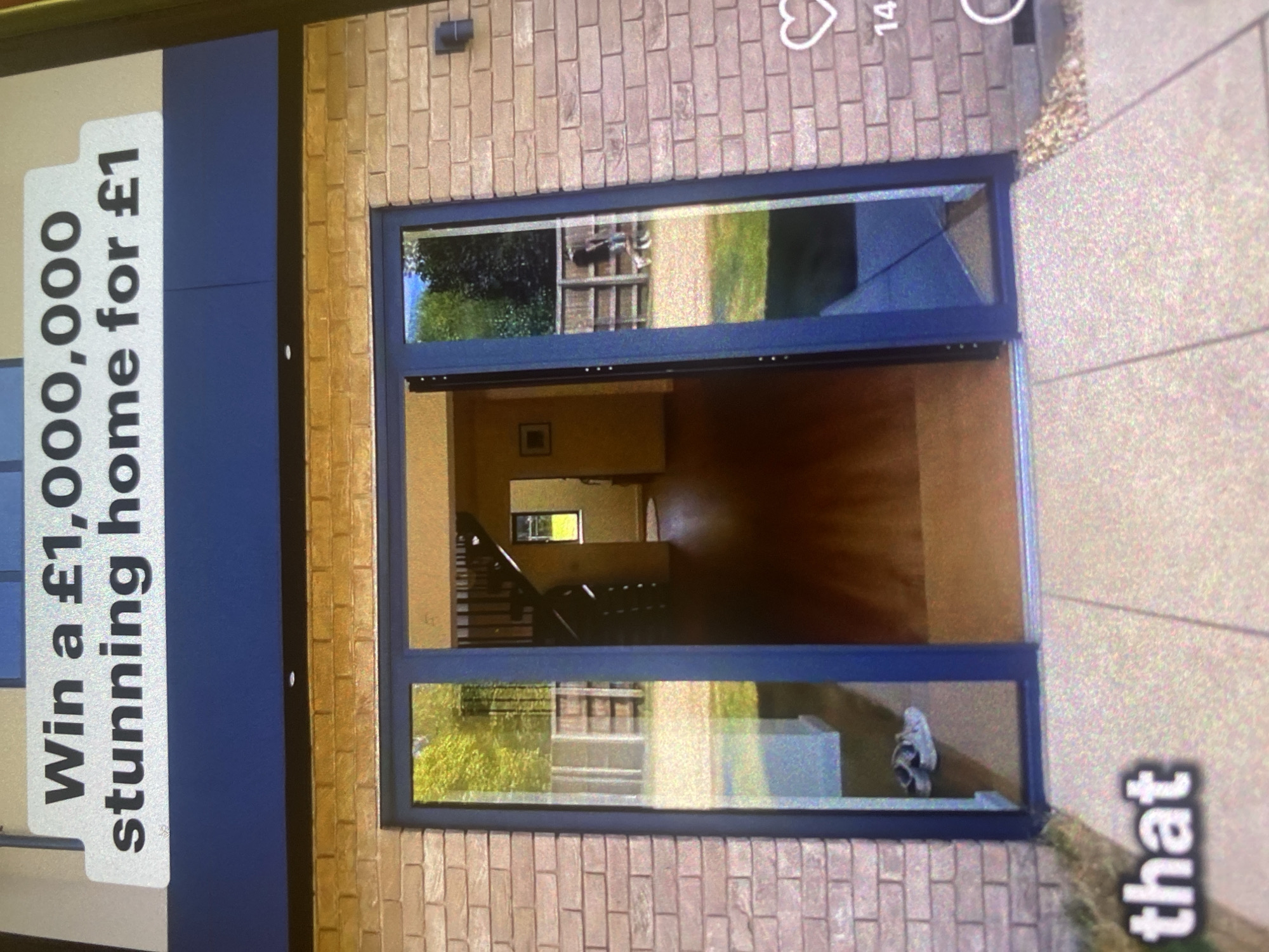 A wide-angle, eye-level exterior shot captures the front entrance of a modern brick house. A large, dark blue framed glass door dominates the center of the frame, revealing an interior hallway with dark wood floors and a spiral staircase. To the left of the door, a pair of white sneakers rests on the interior floor.
Above the doorway, a white banner displays text in black, bold lettering: "Win a £1,000,000 stunning home for £1". The brickwork of the house is a light, sandy brown. The ground in front of the door is paved with a light-colored speckled material, with a border of small, light-colored gravel.
In the background, visible through the glass doors, a person is walking a dog in a grassy backyard enclosed by a wooden fence. To the bottom left of the image, the word "that" is visible in large, white, bold lettering. A heart icon with the number "14" below it is present on the right side of the frame, suggesting social media engagement. The photograph appears to be taken during daylight with natural lighting. No specific location in Bassetlaw, United Kingdom, is identifiable from the image itself.