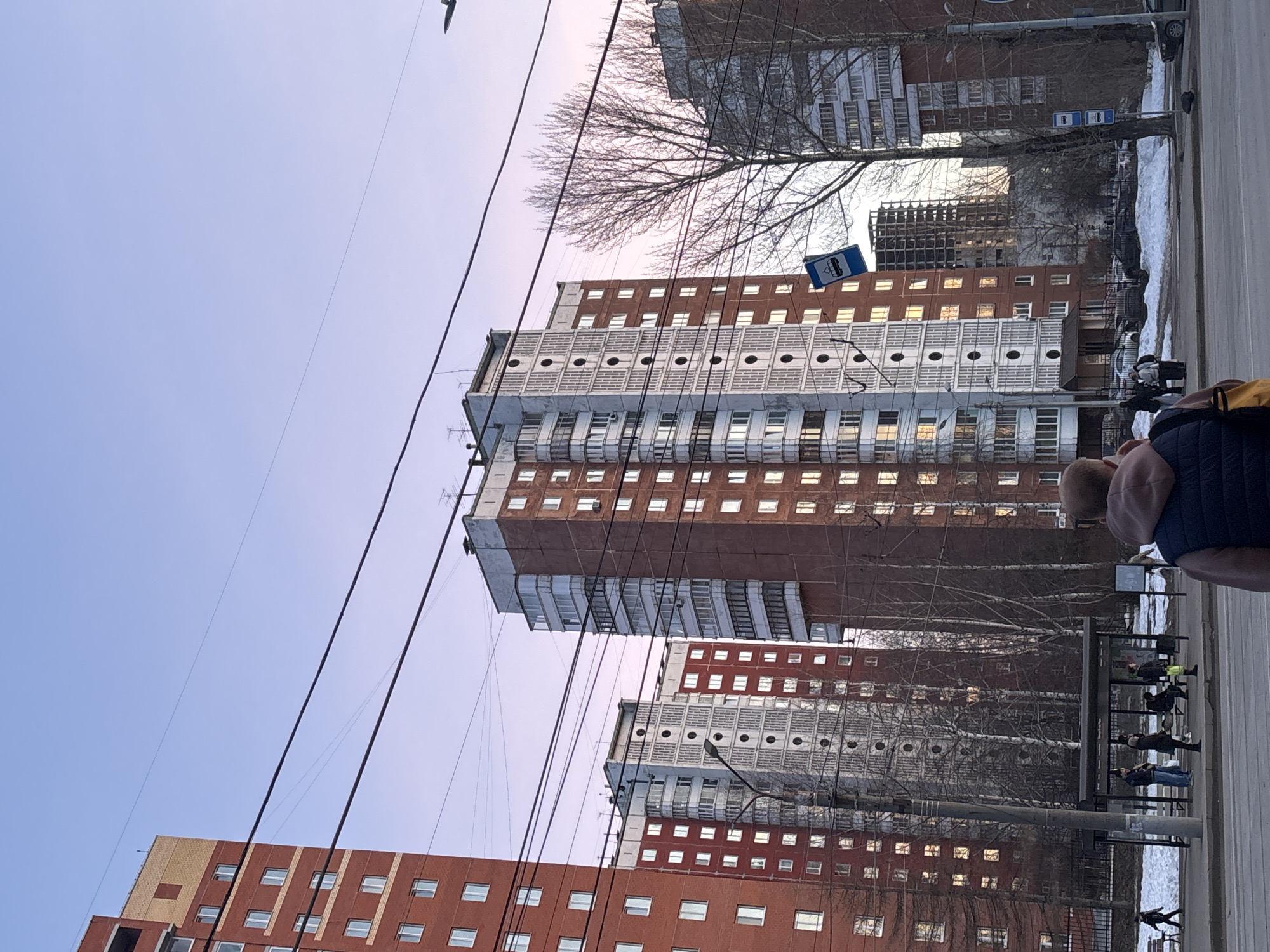 The image shows a view of high-rise apartment buildings in Perm, Russia, taken from street level. The buildings are constructed of red brick and white concrete, with distinctive rounded details on some floors. Several utility wires crisscross the foreground against the sky.
To the right, a person's head and shoulder are visible, suggesting the picture was taken from the perspective of someone on the sidewalk. Further down the street, a bus stop is present with several people waiting at it. Snow is visible on the roadside. There are bare trees between the buildings and the street.