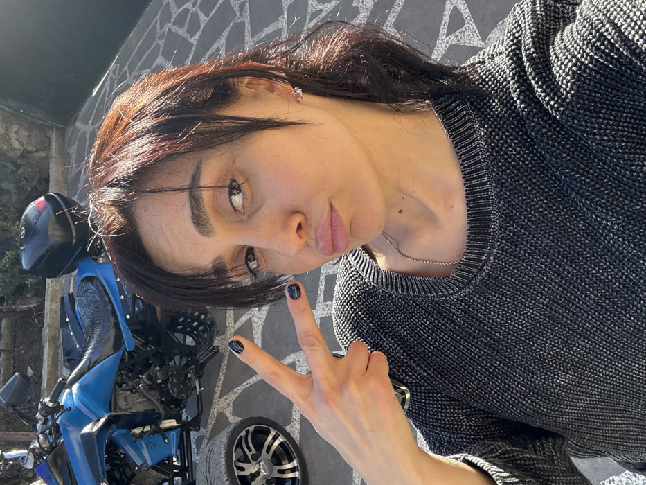 The scene depicts an outdoor setting in Bolnisi, Georgia, under direct sunlight. An adult female is positioned in the foreground, facing the camera for a selfie. She has dark brown hair with bangs, wears a dark gray knitted sweater, a thin silver-colored chain necklace, and small, light-colored stud earrings. Her fingernails are painted black. She is pouting her lips and holds her left hand aloft, forming a peace sign with her index and middle fingers extended. Behind her, partially visible to the left, is a blue all-terrain vehicle (ATV) with a black seat and a black hard-shell storage case mounted on its rear. A portion of the ATV's front left wheel, featuring a silver and black multi-spoke rim, is visible. The ground is paved with irregularly shaped, light-colored stones.