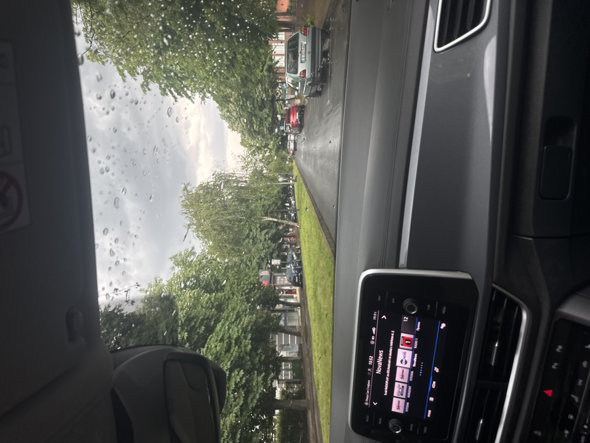 The photograph shows a view from inside a car, looking out onto a street lined with green trees. Raindrops are visible on the windshield, suggesting it is raining. The car is stopped in traffic, with other vehicles visible ahead on the road. The car's dashboard and infotainment system are in focus, showing the time as 15:52 and the radio station as NovaNews. The location is specified on the infotainment screen as “Улица Сан Стефано,” presumably a street name. The city and country are identified by text on the infotainment screen as Unknown and Unknown, respectively.