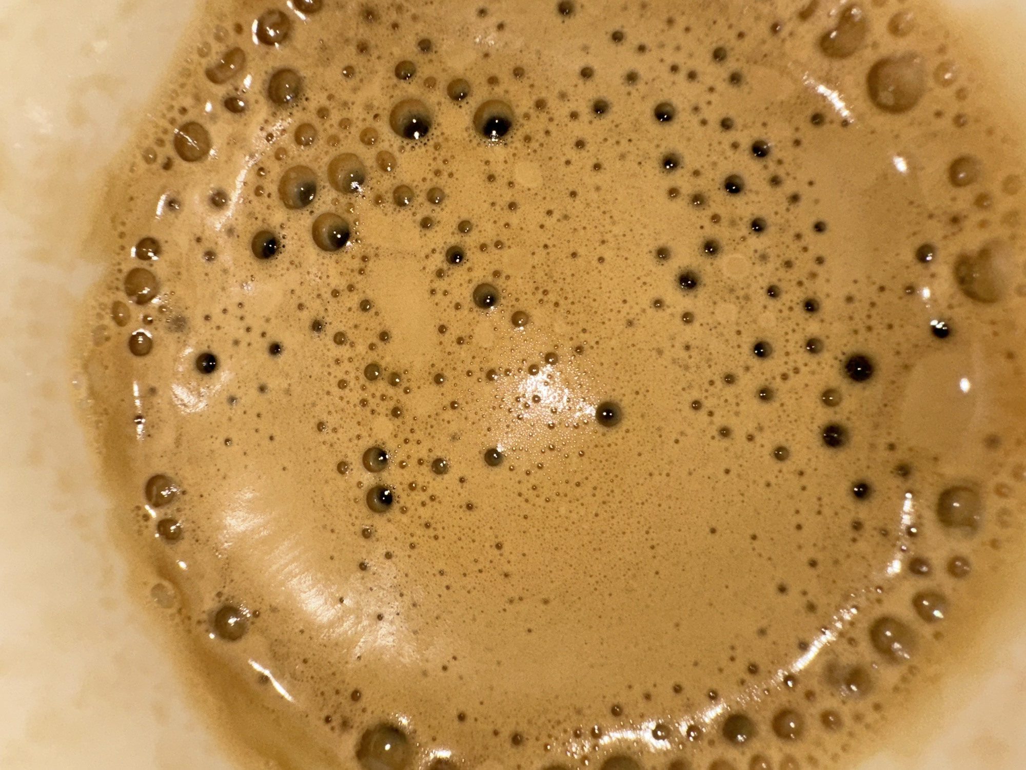 The image is a close-up of the surface of a beverage, likely coffee, with a light brown frothy texture. Numerous small bubbles are visible throughout, with some larger bubbles concentrated along the edges. The background is out of focus and appears to be a light color, possibly the inside of a cup. There is no evidence to suggest the location is Stara Zagora, Bulgaria. There are no people, subjects, objects, actions, interactions or environment visible in the image.