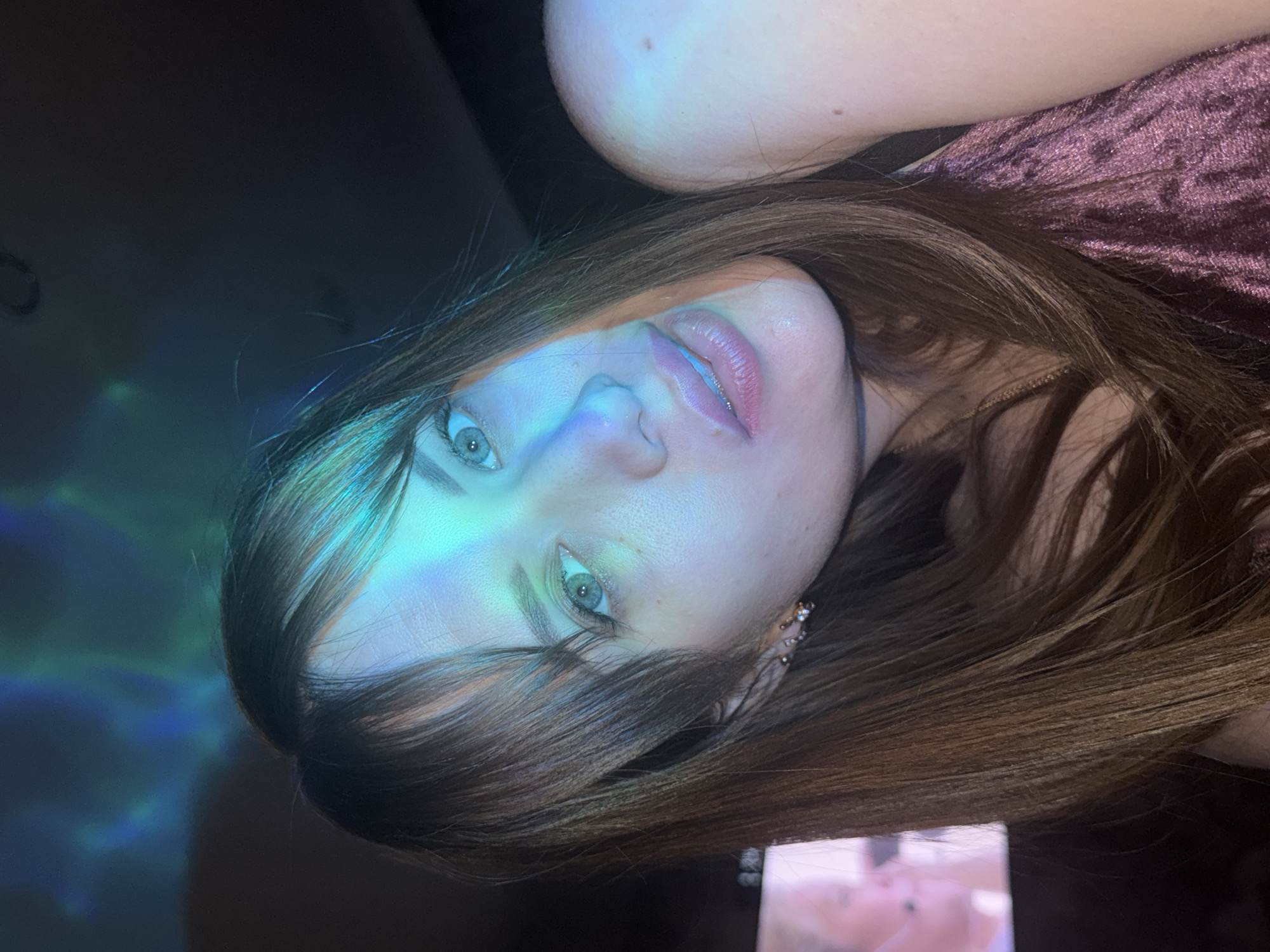 A medium shot shows a fair-skinned woman with light eyes and light brown hair lying down. Her head is positioned in the lower center of the frame, tilted slightly to the right. Her lips are slightly parted, and she seems to be looking at the camera. Her hair is draped over her shoulder, and she wears a mauve-colored dress or top. The woman is possibly wearing earrings in her left ear.
The background is a mix of darkness and light. There's a projection of blue and green light patterns on the left side of the image, while the rest of the background is darker. The specific location within the city of Chaikovsky, Russia, cannot be determined from the image. There are no specific actions or interactions occurring other than the woman posing for the photo.