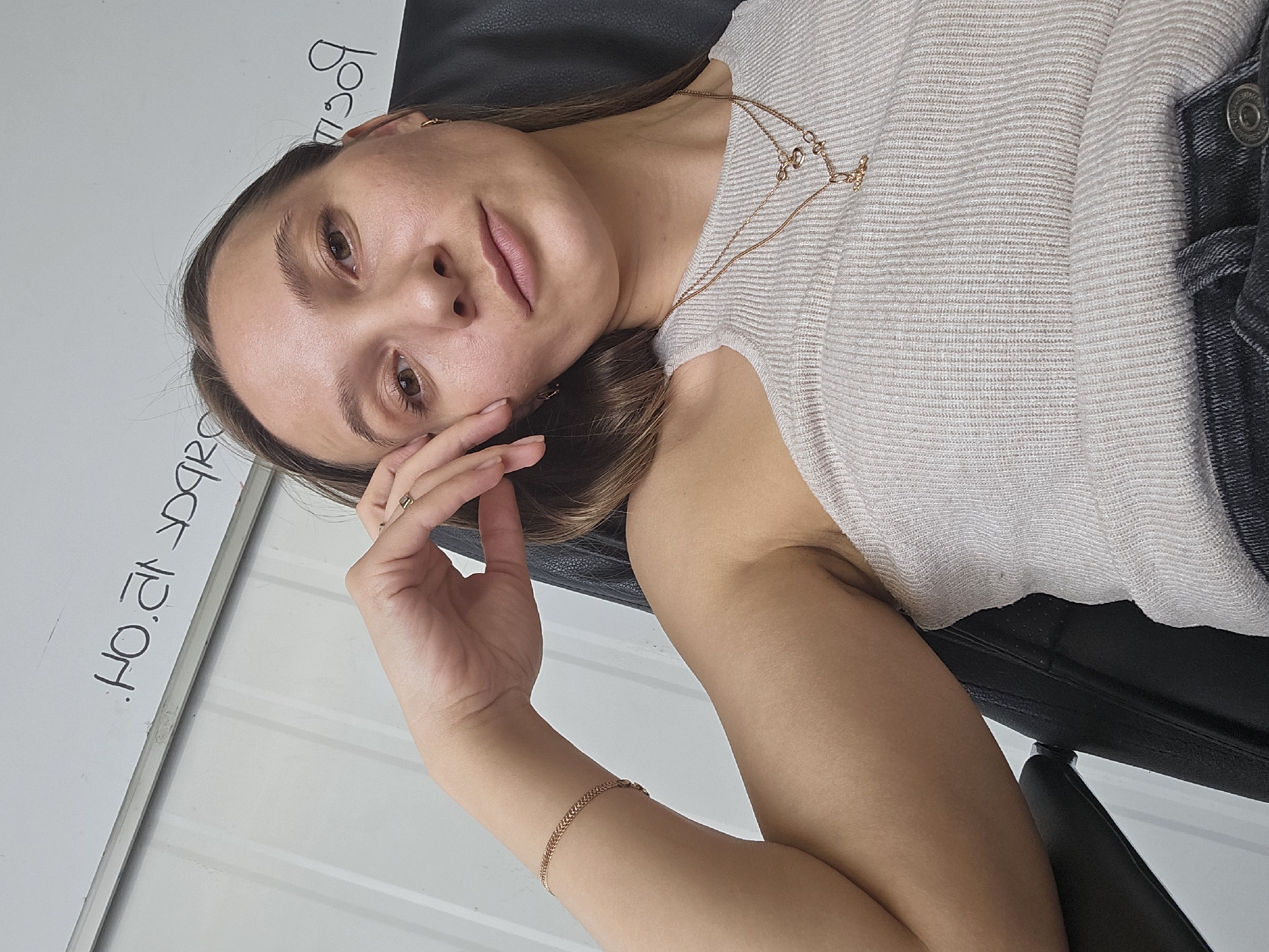 The image displays a close-up, chest-up portrait of a light-skinned female individual, taken indoors in the city of Chaikovsky, Russia. The subject has light brown hair and light brown eyes, with a neutral facial expression as she looks directly at the camera, suggesting a selfie. Her left arm is bent, and her left hand rests against her left cheek and jawline.
She is dressed in a light-colored, possibly off-white, ribbed tank top. Her visible accessories include a gold-toned chain bracelet on her left wrist, a gold-toned band ring on her left ring finger, and small gold-toned hoop earrings in both ears. She wears two layered gold-toned necklaces: one with multiple small heart-shaped charms, and another with a distinct orb-like pendant. Dark-colored fabric, consistent with denim jeans, is visible at the bottom right of the frame.
The individual is seated in a black chair, likely upholstered in leather, with its armrest and a portion of the back visible on the right side. The background consists of a white surface, resembling a whiteboard or dry-erase board. Handwritten text and numerals in black marker are present on this surface, appearing upside down and mirrored. Visible elements include partial Cyrillic characters and numerals such as "НО.ЯГ", "д9q", and "ЯЗОР".