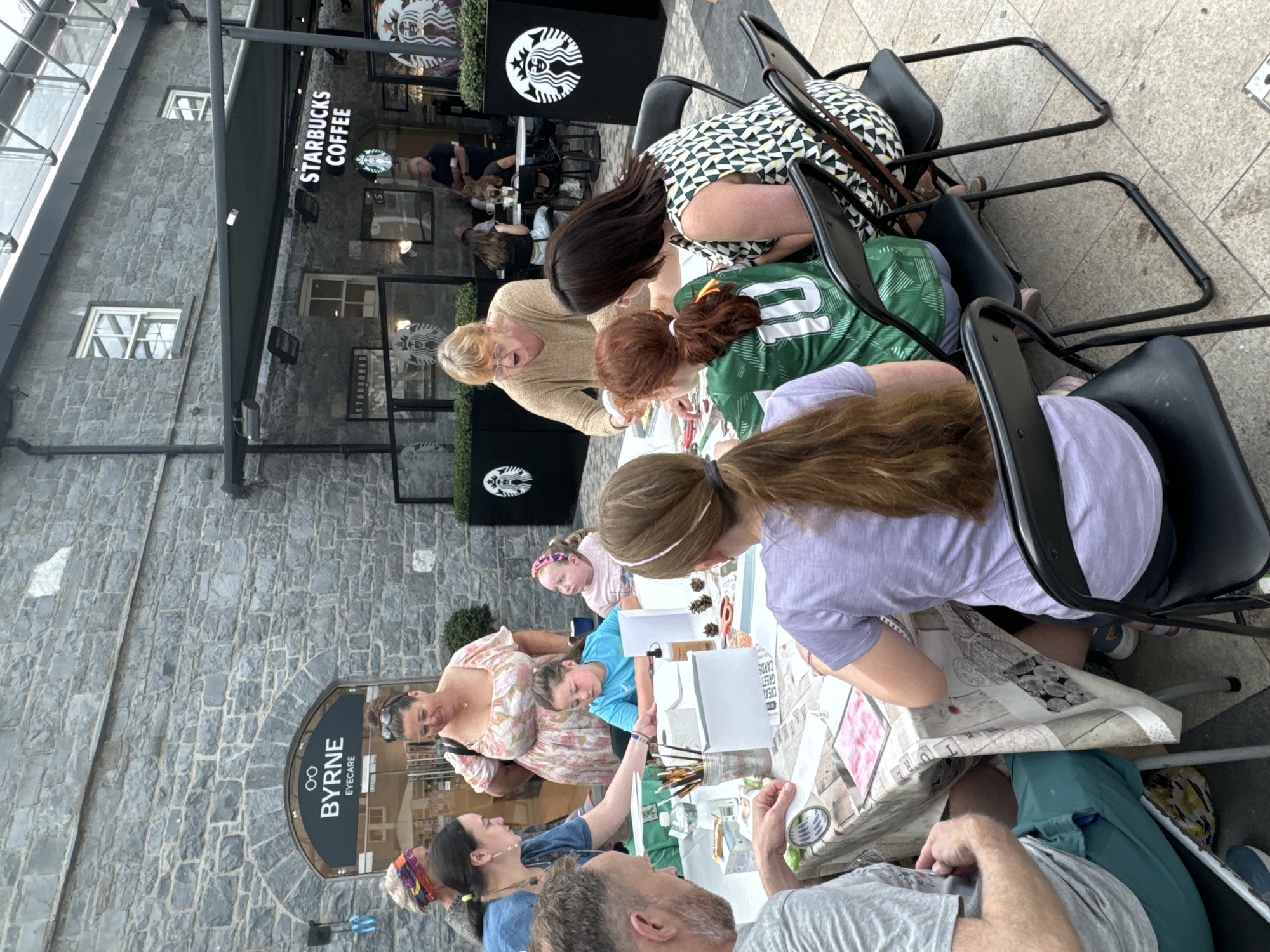 The image captures an outdoor activity at the Kilkenny Design Centre, located in Kilkenny, Ireland. A group of people, including adults and children, are gathered around a table, engaged in a craft or art session. The table is covered with a patterned cloth and laden with art supplies such as paints, brushes, paper, and small decorative items like pinecones.
Several individuals are actively participating: a woman in a blue shirt is guiding a young girl in a turquoise shirt, who is focusing on the materials on the table. A man in a grey t-shirt and teal shorts is also seated at the table, looking at something in his hands. Another woman, wearing a patterned dress, is leaning in to assist a child in a green soccer jersey with the number 10 on the back. A woman in a beige top appears to be instructing or explaining something. In the background, other patrons can be seen in a Starbucks cafe.
The setting features a stone building facade with a visible "BYRNE EYECARE" sign above an arched entrance. Adjacent to this, a Starbucks cafe with its iconic green branding is present. The ground is paved with light-colored tiles. The overall atmosphere suggests a community event or a recreational activity taking place in a public space within the city.