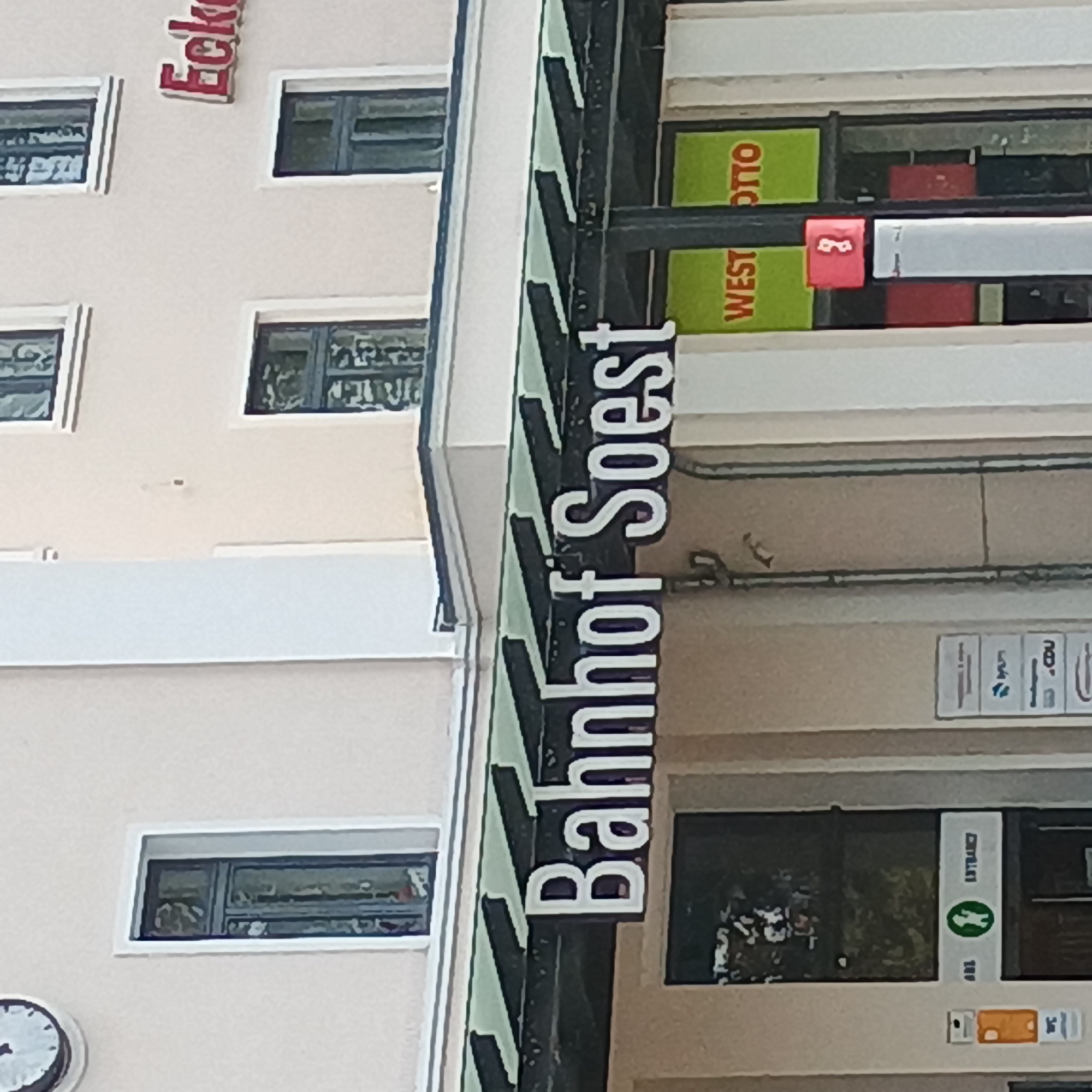 The image shows the exterior of a building, identified by a prominent sign as "Bahnhof Soest". This indicates it is the train station in the city of Soest, Germany. The building has a light-colored facade with several windows. A clock is visible on the top left of the building, and red lettering that appears to say "Eck..." is visible on the upper right. Below the "Bahnhof Soest" sign, there are storefronts, one of which has green signage with red text reading "WEST OTTO". There is also a red sign with a white number "3" visible. The lower left of the image shows an entrance with a sign featuring a green circle with a white pictogram of a person, and text that reads "ENTRANCE". There are also smaller signs with icons for WC. The scene appears to be during the daytime, with natural light illuminating the building.