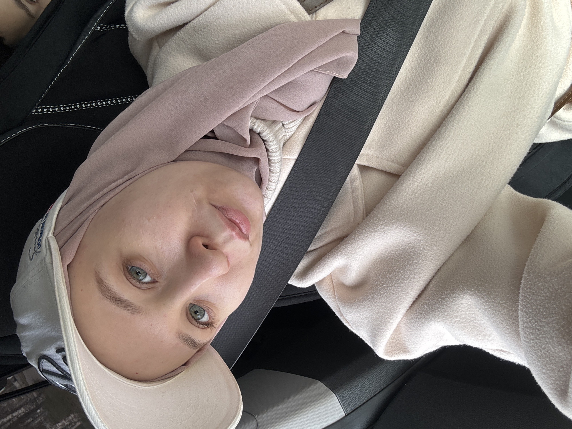 A woman wearing a hijab and a baseball cap is shown from a slightly high angle. She is wearing a seatbelt. The location is not identifiable as Chaikovsky, Russia, without additional visual or contextual information. The background shows interior car elements such as the seat and dashboard.