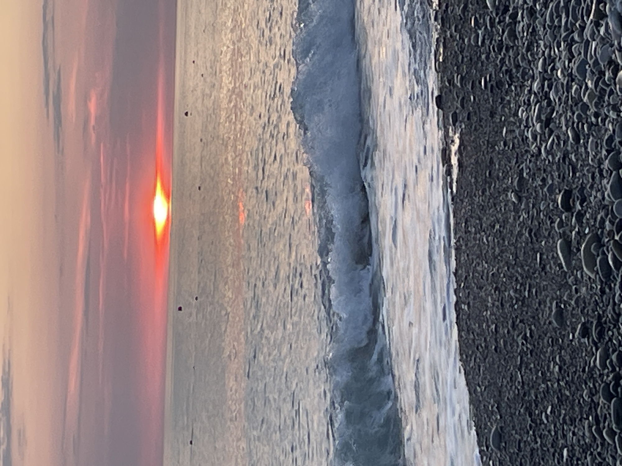 The image depicts a serene beach scene at sunset. The sky is painted with hues of pink and orange, with the sun a bright orange orb just above the horizon, casting a warm glow on the water. The ocean reflects the sky's colors, creating a shimmering effect on the gentle waves. In the foreground, a dark, pebbly beach stretches out, with the waves softly lapping at the shore. A few small, dark buoys are scattered in the distance on the calm sea. The overall impression is one of tranquility and natural beauty. The location is Unknown, Unknown.