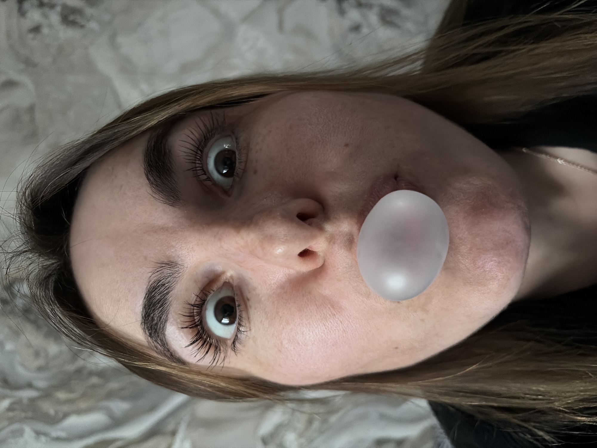 A close-up, slightly distorted, and rotated image shows a light-skinned individual with brown hair blowing a bubble with pink bubblegum. The person has noticeable freckles. The background appears to be a soft, textured surface. There is no evidence to suggest a location in Izhevsk, Russia.
