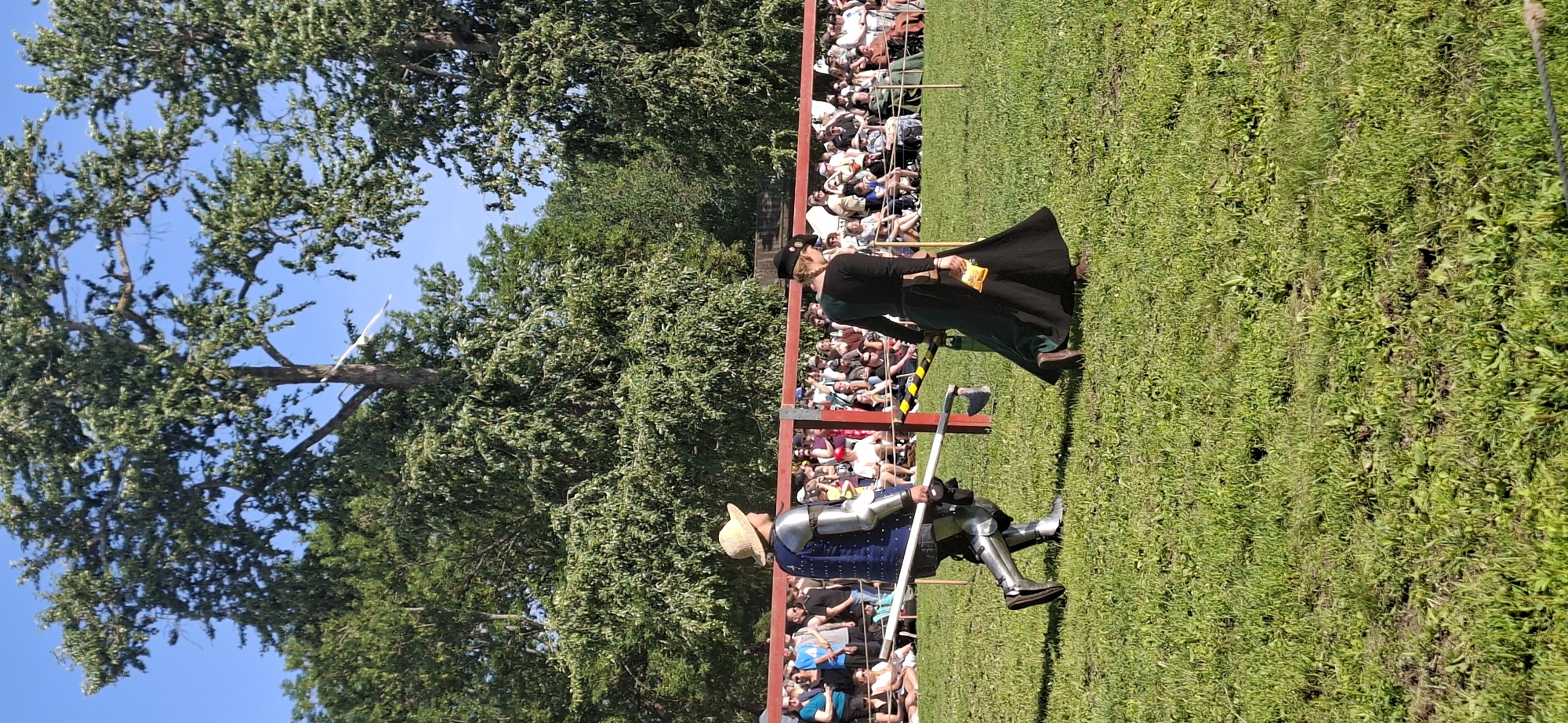 In Turku, Finland, a medieval reenactment event is underway on a sunny day. The scene depicts two individuals in period attire on a grassy field, with a crowd of spectators in the background.
The person on the left is dressed in full plate armor, including a helmet, gauntlets, and greaves. They are also wearing a straw hat over their armor. They are carrying a long wooden staff. This individual is walking to the left, facing away from the camera.
The person on the right is wearing a long, dark green and black dress with a matching head covering. They are carrying a small, yellowish pouch in their right hand. This individual is walking to the right, also facing away from the camera.
A red barrier runs across the background, separating the performance area from the audience. Spectators are seated and standing behind this barrier, watching the event. Trees with lush green foliage form a backdrop, and the sky is a clear blue.
The overall atmosphere suggests a historical reenactment or festival, likely in an outdoor public space in Turku.