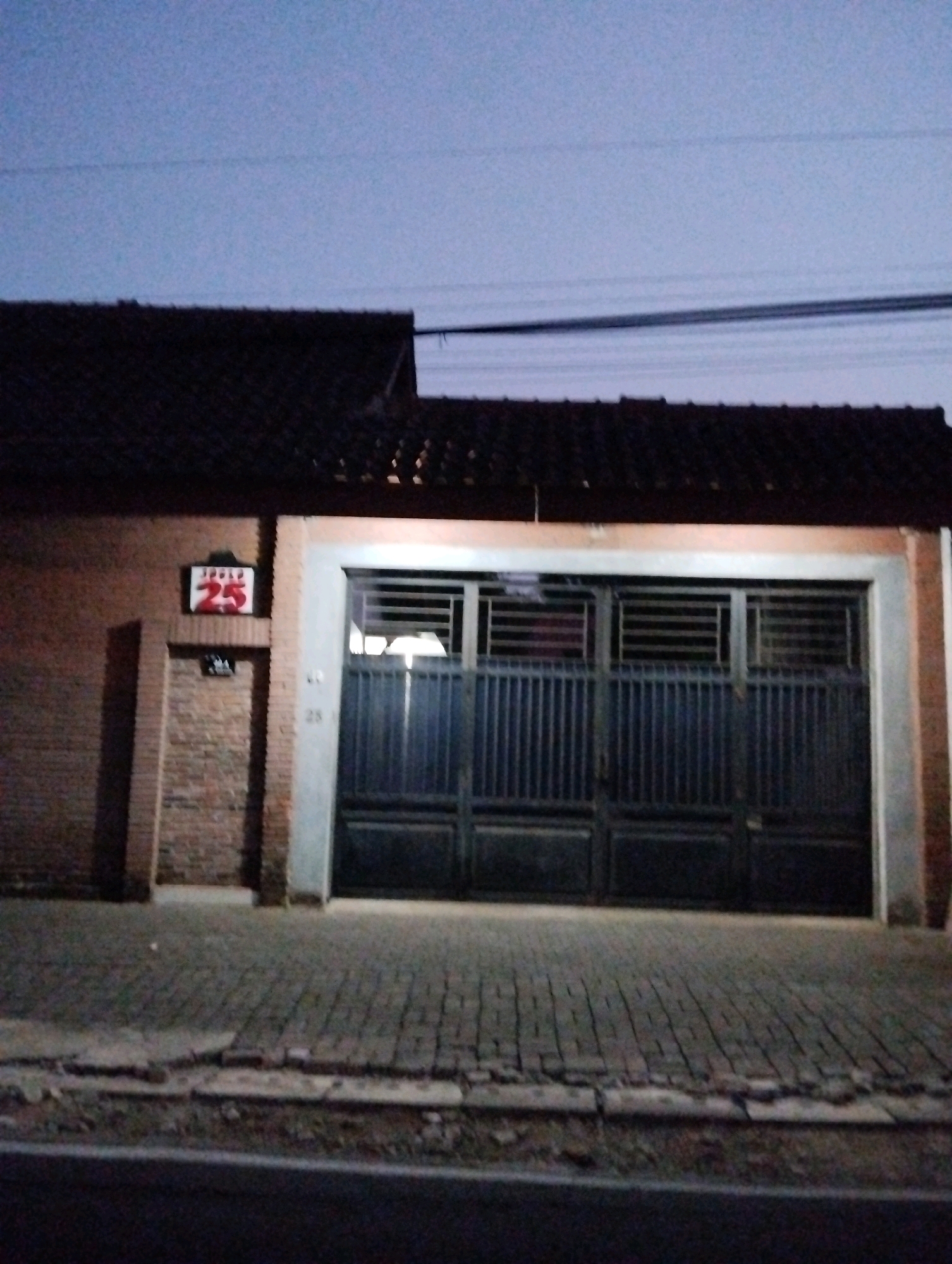 The image shows the exterior of a building at night, likely in Salatiga, Indonesia. The building has a dark tiled roof and brick walls. A large, dark metal gate is the central feature, with horizontal slats and vertical bars. To the left of the gate, a brick wall has a sign with the number "25" in red digits within a white rectangular frame. Below this sign is a smaller, unreadable sign. A narrow brick doorway is visible to the left of the main gate. The ground in front of the building is paved with bricks, and a street is visible in the foreground. The sky above is a dim blue, indicating either dusk or dawn. There are no people visible in the image. The overall impression is that of a residential or commercial property entrance.