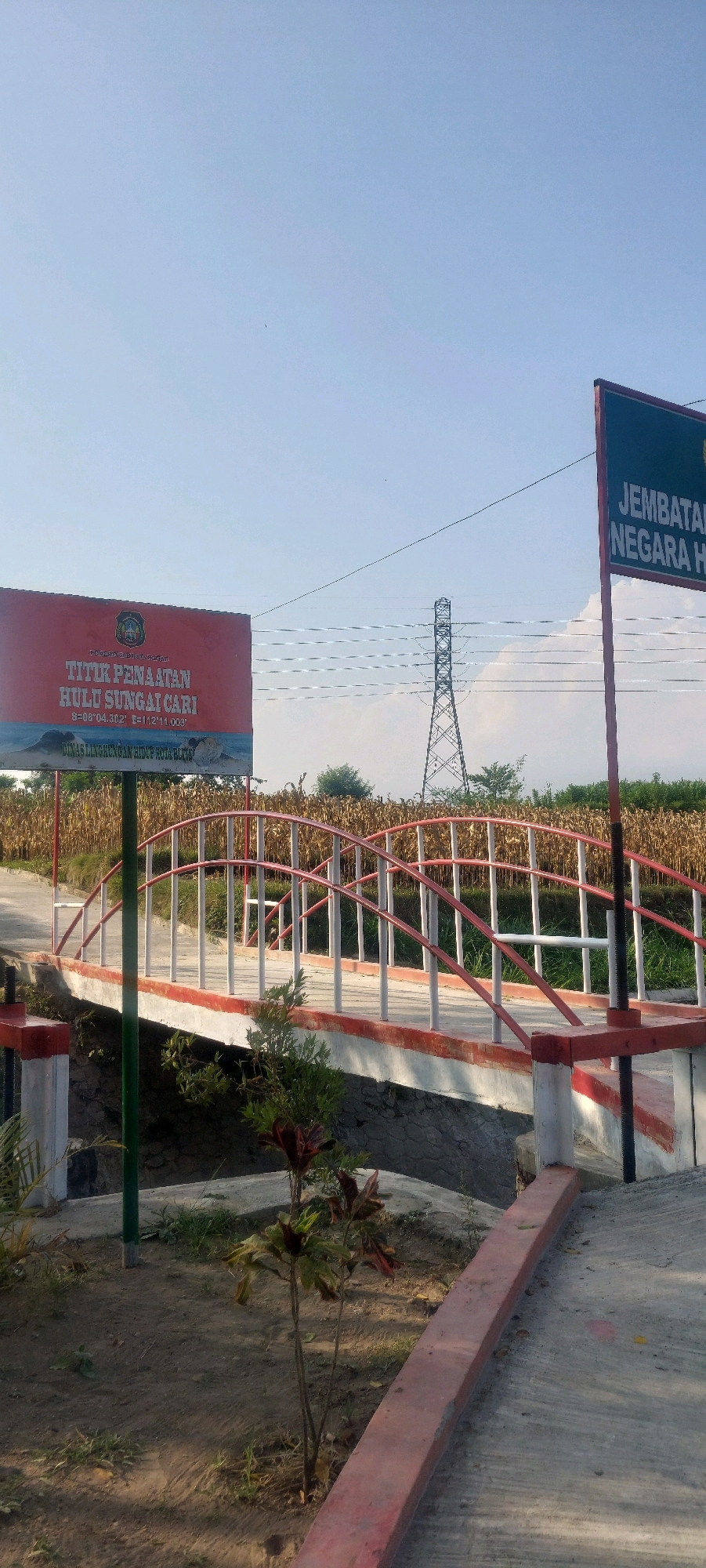 An outdoor, eye-level, vertical shot shows a bright, sunny day with a clear blue sky dominating the upper half of the frame. In the foreground, a concrete pathway with a red curb leads the eye towards a small bridge with white vertical posts and red arched railings. The bridge spans a dry, rocky streambed. Beyond the bridge, a field of dry corn stalks stretches across the middle ground. To the left of the bridge, a red sign with Indonesian text and an emblem is mounted on a green pole. To the right, another sign with red lettering against a dark background is partially visible. In the background, a tall, lattice-like electrical tower stands against the sky. The overall impression is of a rural or semi-rural location.