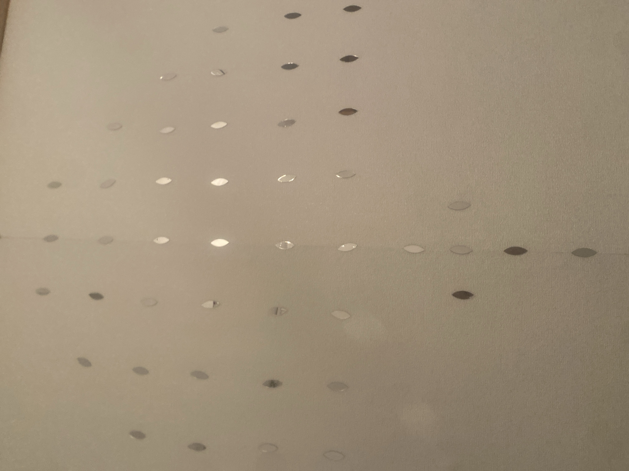 The image displays a patterned background with multiple small, reflective oval shapes. These shapes are scattered across the surface in a seemingly random, yet somewhat organized, fashion. The background itself is a muted, neutral color, possibly a light beige or cream, with a subtle texture. The reflective elements are varied in their orientation and in the way they catch the light, creating small glints and highlights. There are no people, animals, or any other distinct objects visible in the image. The scene does not provide any information that would allow for a specific location within Boraldai, Kazakhstan to be identified.