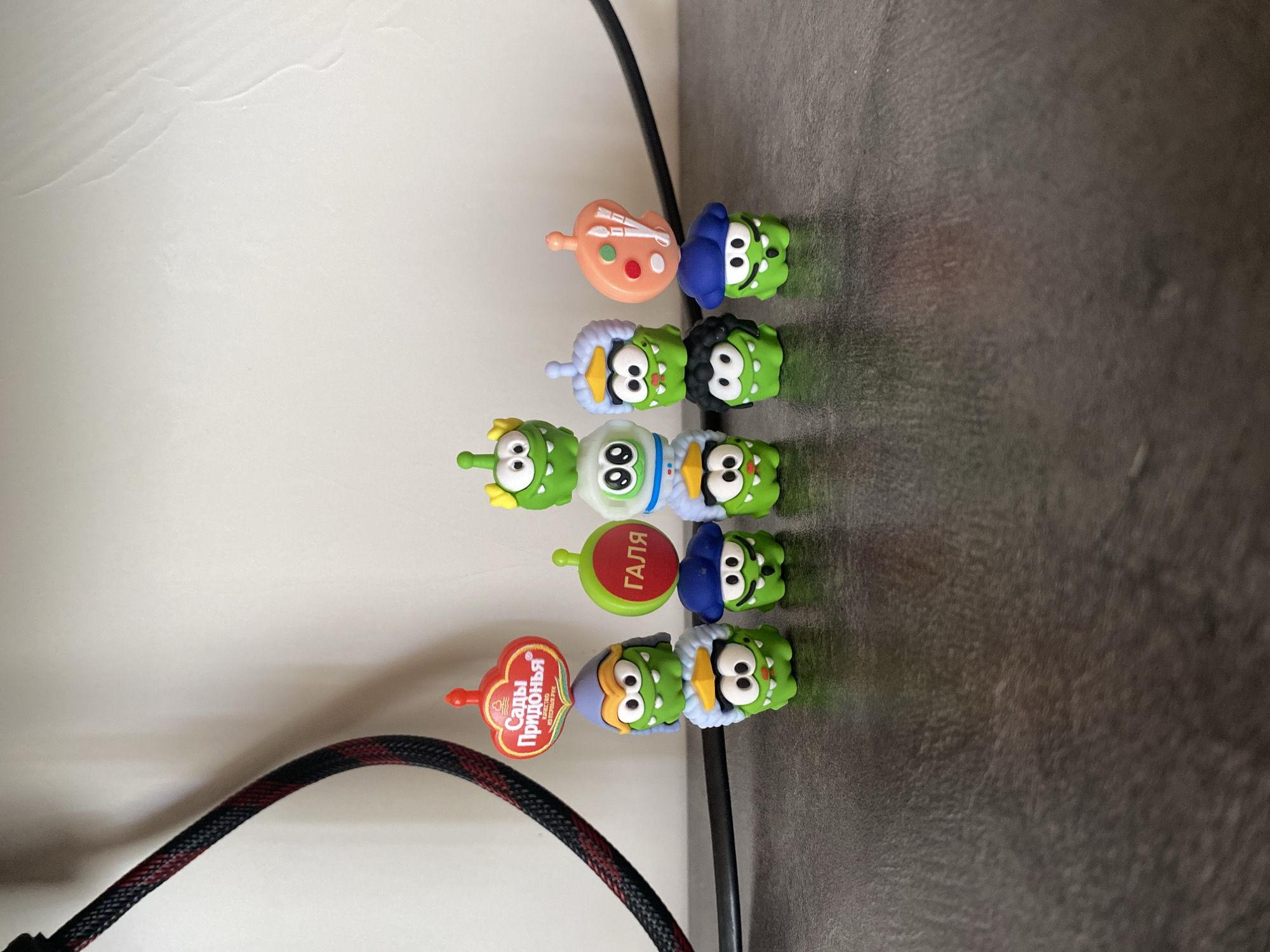 The image shows a collection of ten small, green, cartoon-like figures, resembling characters from the game "Cut the Rope," arranged on a dark, textured surface, possibly a table. The figures are of various heights and some are stacked. They have large eyes and mouths, and some are adorned with hats or other accessories, such as a blue beret, a white astronaut helmet, a light purple helmet, and a black wig. One figure is topped with a small, round, red object with Russian text that translates to "Sadovye Pridonya." Another figure has a red circle with the Russian word "GALA" on it. A third figure is topped with a pink artist's palette with paintbrushes and dabs of paint. In the background, a white wall is visible, and to the left, a black and red braided cable is partially visible. The scene is lit from the front, casting minimal shadows. The location in Kisliakovskaya, Russia, is not discernible from the visual evidence in the image.