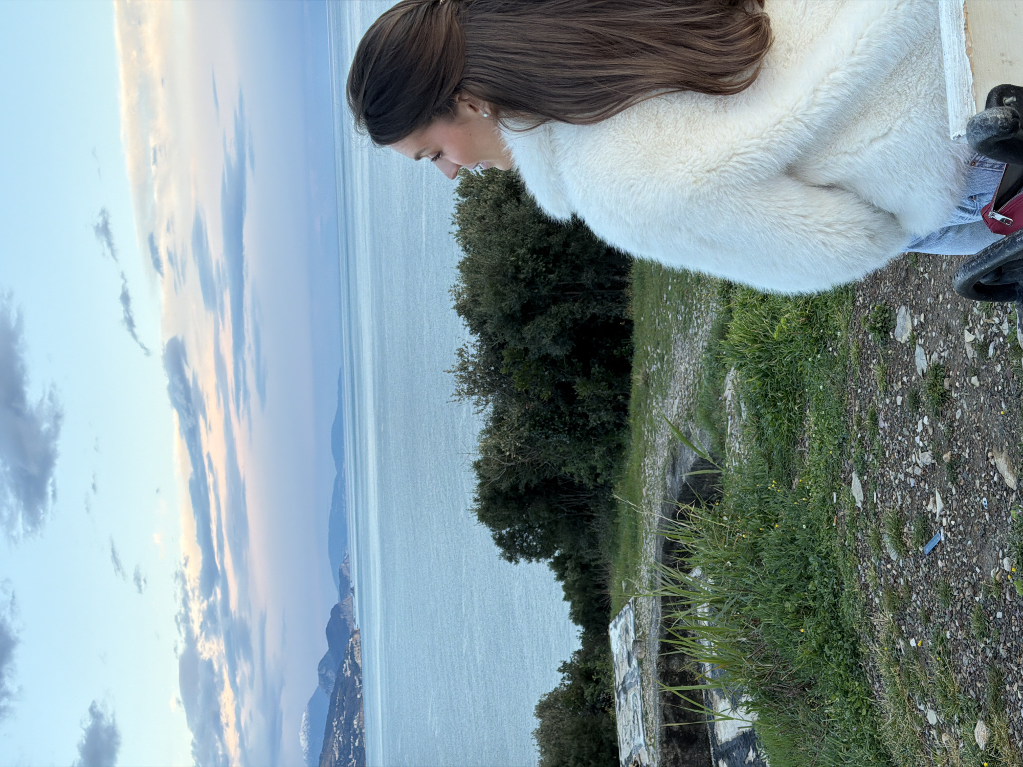 A young woman in a white furry coat is seen from the waist up, looking out at a serene sea and sky. The sky, on the left of the image, transitions from a soft blue to light pink with scattered clouds. The sea, spanning horizontally, reflects the sky's colors. Lush greenery and trees grow on what appears to be a clifftop to the right, with a stone wall partially visible. The foreground consists of gravel and grass. The location is possibly near Budva, Montenegro, given the coastal setting and typical Montenegrin landscape.