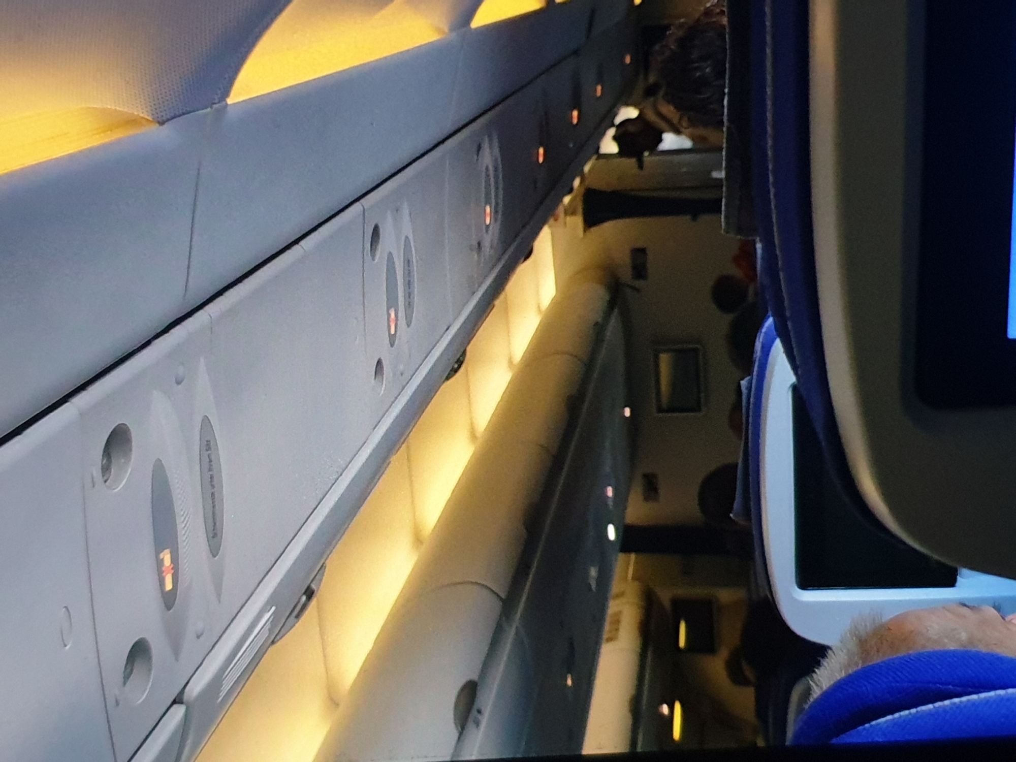 The image shows the interior of an airplane cabin. Overhead, there are overhead bins with lights and passenger control panels. The passenger control panels indicate the "fasten seatbelt" sign is illuminated. The seats appear to be occupied by passengers. One passenger is visible on the right side of the frame wearing a blue neck pillow and partially obscured by the seat-back entertainment screen.