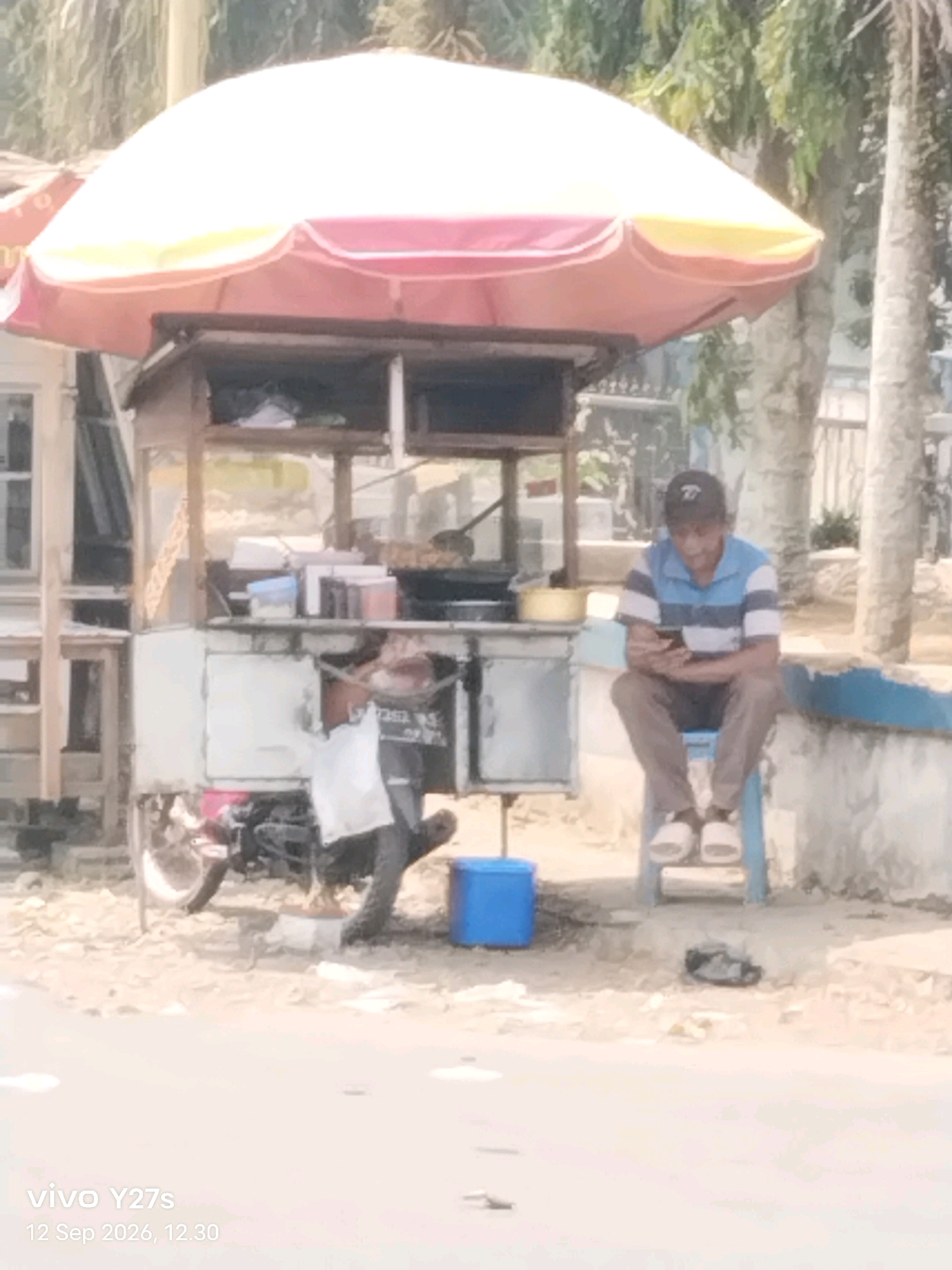 A man sits on a blue plastic stool outside a food stall in Trimulyo, Indonesia, looking at his phone. The food stall, covered by a red and white umbrella, has a white base and wooden shelves displaying various items. To the left of the stall, a motorcycle is parked. A blue plastic container sits on the ground near the stall. The man wears a striped blue and white shirt, brown pants, a black baseball cap, and white slip-on shoes. He is positioned on the right side of the frame. The background features trees and a low wall. The ground is covered in loose gravel and dirt. The overall lighting suggests it is daytime.