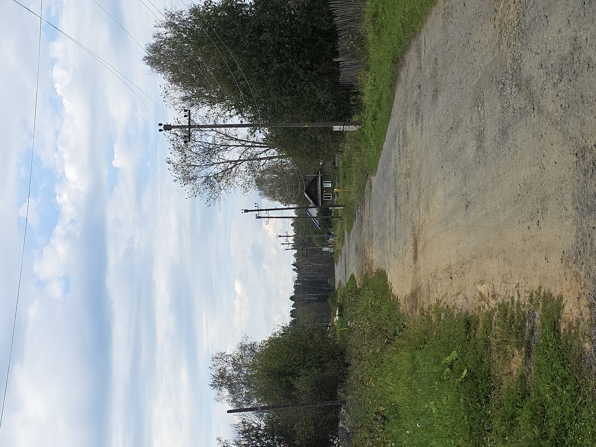 The image depicts a rural road in the city of Magnitka, Russia. The road surface is a mix of dirt and gravel, with several potholes visible. On either side of the road, there is tall grass and vegetation. Power lines and utility poles are present along the road. In the distance, a small wooden house can be seen nestled among trees. The sky is partly cloudy, with a mix of blue sky and white clouds. The overall scene suggests a quiet, undeveloped area.