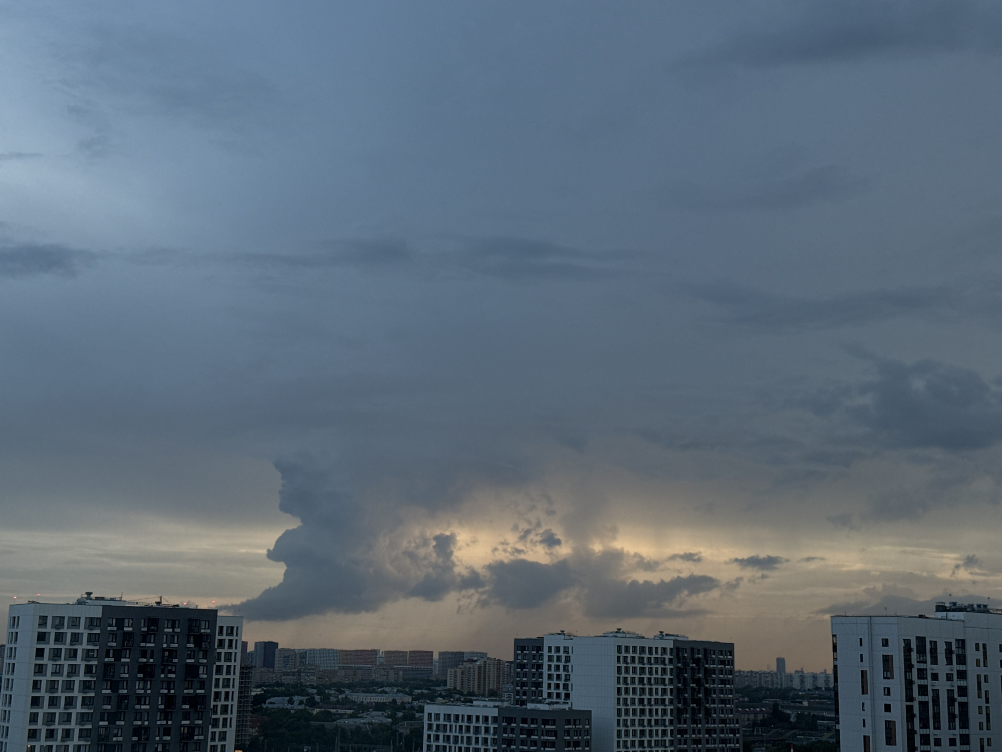 The image shows a cloudy sky over the city of Lyubertsy, Russia, with several apartment buildings in the foreground. The sky is dominated by dark, heavy clouds, with a patch of lighter, yellowish light breaking through in the center. A prominent, towering cumulonimbus cloud formation is visible on the left side of the image, its shape somewhat resembling a figure. The apartment buildings are modern, multi-story structures with numerous windows. In the background, a dense urban landscape with more buildings and some greenery can be seen stretching towards the horizon. The overall atmosphere is somber, indicative of an approaching storm or overcast weather.
