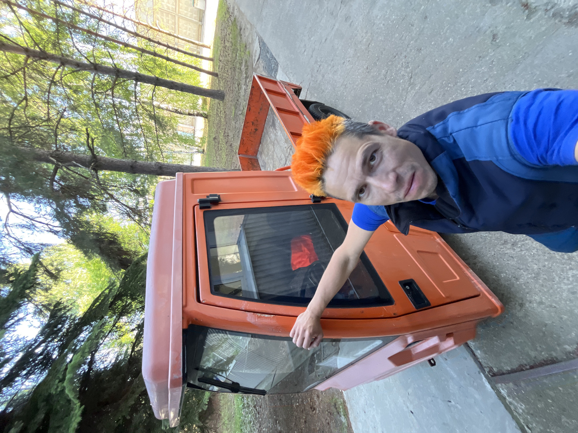 A man with bright orange hair is standing next to an orange utility vehicle. The man is wearing a blue and black vest over a blue shirt. His right arm is extended and his hand is resting on the side of the vehicle's cabin. The vehicle has a small cargo bed and is parked on a concrete surface. In the background, there are trees and a fence. The scene appears to be outdoors in an urban or semi-urban area, possibly in Chaikovsky, Russia, given the context. The lighting suggests it is daytime, with sunlight filtering through the trees.