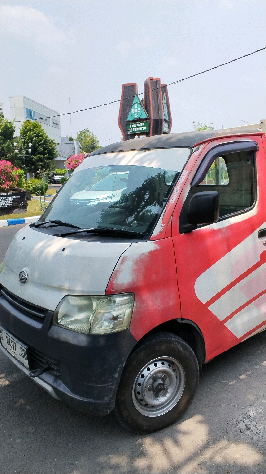 The image captures a red and white Daihatsu van parked on an asphalt road in Banjaragung, Indonesia. The van's hood is white with red paint chipping, and its bumper is dark gray. A license plate reading "S 8497 SB" is visible. In the background, a sign with "UKS" and "KABUPATEN MOJOKERTO" is prominently displayed, along with a modern white building and lush green trees. The sky is bright and clear. The overall scene is an outdoor urban environment.