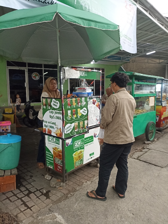 A street food vendor is selling Exo Tea in Jatinangor, Indonesia (latitude: -6.935, longitude: 107.773). The vendor is a woman wearing a headscarf. A man is purchasing Exo Tea. The food cart has a large green canopy and the vendor is standing behind it, showing the Exo Tea products to the customer. Behind the food cart, another street vendor is seen selling various items from a green cart with a canopy, standing on the same street. The vendor is wearing a tan jacket and blue jeans. The street is paved with gray concrete and the background is a building with a green door. The vendor's food cart is displaying a variety of drinks, including chocolate series and coffee series. The image is taken during the day.
