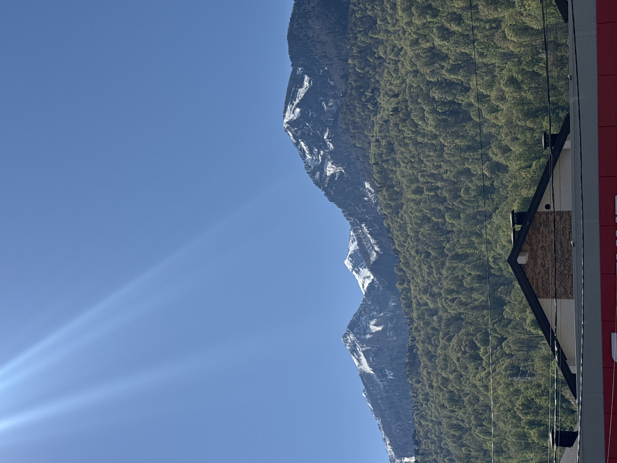 The image shows a mountain range partially covered in snow and dense green trees, juxtaposed against a clear blue sky with visible sun rays. In the foreground, the edge of a building is visible, featuring a roof with dark trim and brick siding, as well as red siding further on. The setting is likely Krasnaya Polyana, Russia, known for its mountainous terrain and ski resorts. There are no people present in the image. Power lines can be seen running across the tree line and roof.