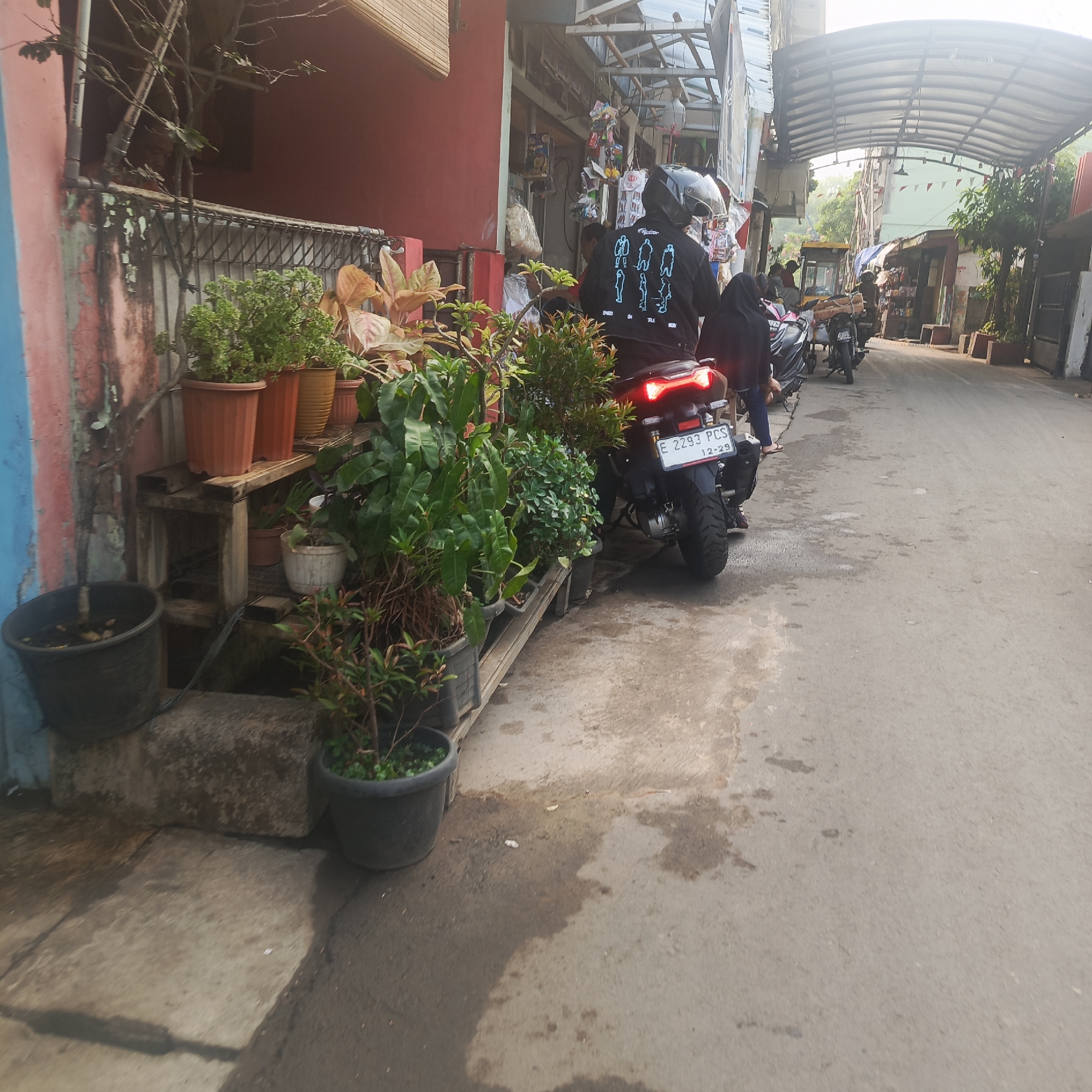 The image depicts a street scene in South Jakarta, Indonesia, likely in a residential or mixed-use area. In the foreground, a person wearing a black jacket with a distinctive white graphic pattern and a helmet is seated on a motorcycle. The motorcycle is parked on the side of a paved road. Adjacent to the motorcycle, on the left side of the frame, are several potted plants, including leafy green varieties and some with reddish-brown leaves, placed on shelves and on the ground. The architecture visible includes buildings with reddish-pink and light-colored facades, some with metal grating and awnings. Further down the street, other motorcycles and a small yellow cart or kiosk can be seen. The overall atmosphere suggests a typical day in a bustling urban environment.
