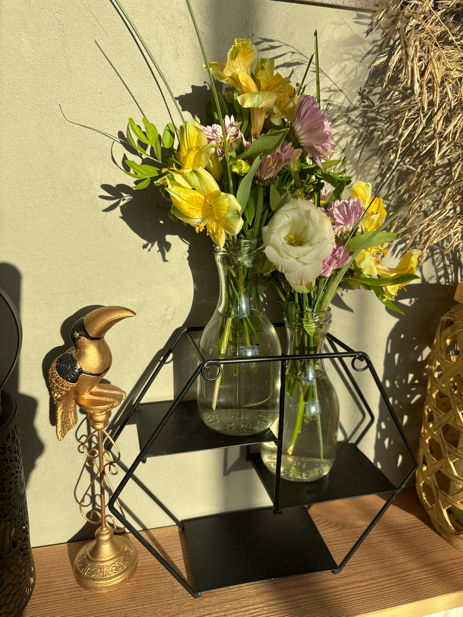 In Sapronovo, Russia, a still life is depicted on a wooden shelf. Two glass vases containing a bouquet of yellow and pink flowers with green foliage are placed on a black geometric shelf. To the left of the vases, a golden toucan figurine stands on an ornate golden base. To the right of the bouquet, a woven basket-like object is visible. The background is a textured wall with shadows cast by the objects and sunlight.