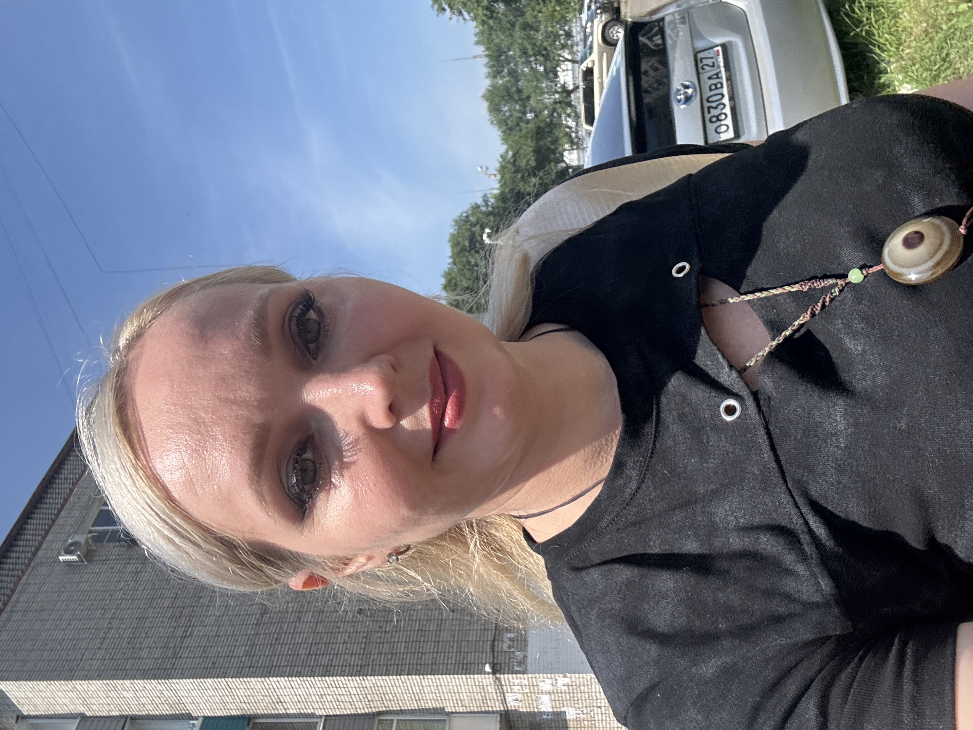 A medium shot captures a woman with blonde hair, fair skin, and green eyes. She is wearing a black t-shirt with cutouts and a necklace with a large, patterned pendant. Her makeup is visible, including eyeliner and lipstick. The background includes a brick building, trees, and a silver car with a license plate reading "0830 BA 27". The sky is blue and clear, suggesting a sunny day. The scene appears to be an outdoor urban environment in Komsomolsk-on-Amur, Russia.