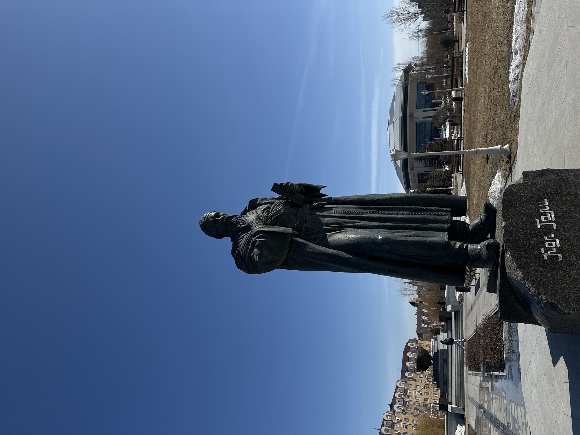 A bronze statue of an older man in a long robe, holding a book, stands in Kazan, Russia. The statue is situated on a stone pedestal with writing in a Cyrillic script. Behind the statue, a cityscape with buildings, trees, benches, and a modern-style building are visible under a clear blue sky. There is some patchy snow on the ground.