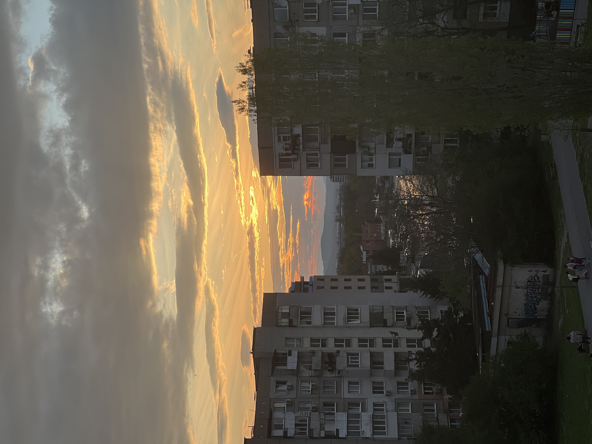 This media file presents an urban landscape in Sofia, Bulgaria, captured during either a vibrant sunset or sunrise. The sky dominates the upper half of the frame, featuring a dynamic display of streaky clouds illuminated in brilliant shades of orange, yellow, and fiery red, contrasting with softer grey and light blue areas. The sun's intense light, partially hidden, creates a glowing horizon that extends across the scene, hinting at the end or beginning of a day.
Below the dramatic sky, several multi-story residential buildings, characteristic of urban architecture in the region, occupy the mid-ground and lower sections. These grey structures feature numerous windows and balconies, with some showing external air conditioning units. Interspersed among the buildings are trees with fresh green leaves, suggesting the season is spring or early summer.
In the distance, a hazy urban sprawl leads to the faint silhouettes of a mountain range, visible beneath the colorful horizon. The lower right portion of the image reveals a paved pathway or road bordered by grassy areas and more trees. On a low-rise building or wall near the path, some graffiti with Cyrillic script is faintly visible. A few small figures, appearing to be people, can be seen on the pathway, engaged in casual activity like walking. The scene evokes a sense of calm, with the everyday urban environment transformed by the stunning natural light.