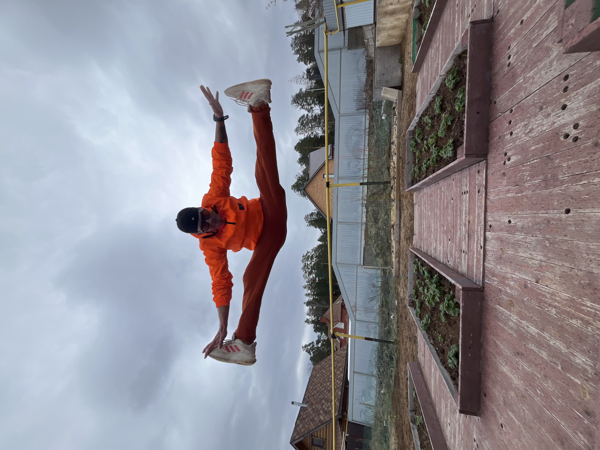 An individual performs a dynamic acrobatic or yoga-like balancing pose outdoors under an overcast sky. The person, who appears male, is wearing an orange long-sleeved jacket, reddish-brown pants, a black baseball cap, and white athletic shoes with three red stripes. A smartwatch is visible on their left wrist. They are captured mid-action, with one leg extended high upwards and one arm reaching towards the sky, while the other leg and arm extend downwards, creating a striking diagonal line across the frame.
The background, located in Chaikovsky, Russia, features a rustic outdoor setting. To the person's right, there are several raised garden beds constructed from weathered, reddish-brown wooden planks. These beds contain dark soil from which numerous small green plants are sprouting. The wooden planks exhibit visible screw or nail holes. In the middle ground, a metal fence separates the immediate area from a line of trees, likely conifers. Further back, various structures are visible, including a house with a brown shingle roof and another building with corrugated metal siding. Yellow gas pipes traverse the scene horizontally. The sky is uniformly grey and cloudy, indicating an overcast day. No discernible text is clearly visible on the clothing or in the surroundings, apart from the three red stripes on the athletic shoes.