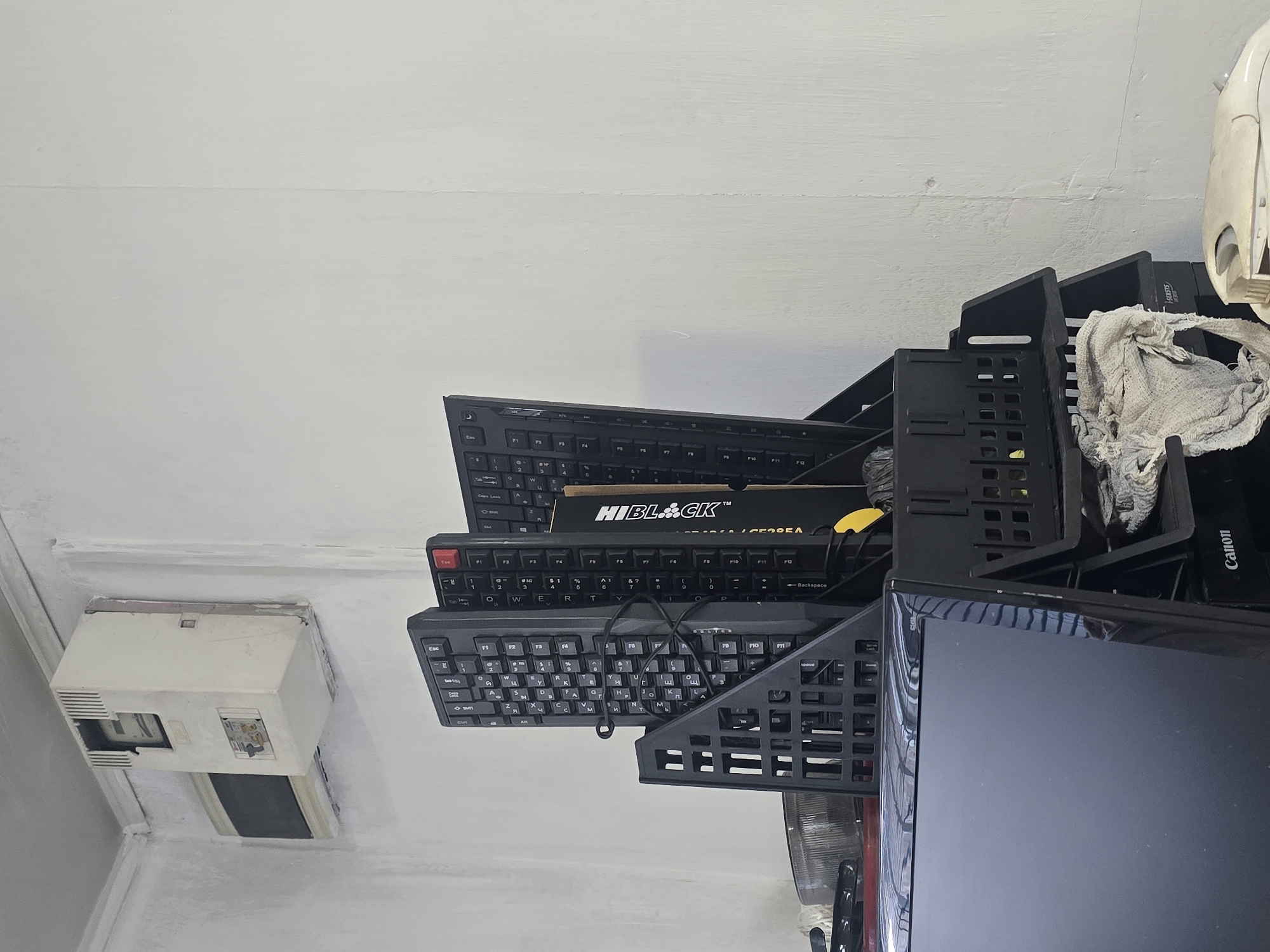 An indoor scene shows a cluttered corner with a white wall and a white ceiling. Mounted on the wall is an old, beige electrical unit with an open compartment showing internal components. Below this unit, and slightly to the left, is a small, dark-colored vent or window. In the foreground, a black computer monitor is visible, partially obscured by a black plastic organizer holding several keyboards. One keyboard is Cyrillic-labeled. A black object, possibly a car headlight, rests on a red surface to the left of the keyboards. A piece of torn, light-colored fabric is draped over the plastic organizer. A white computer mouse is situated on the floor in the lower right corner of the image. The scene appears to be in a storage area or workshop. The location is not specified.