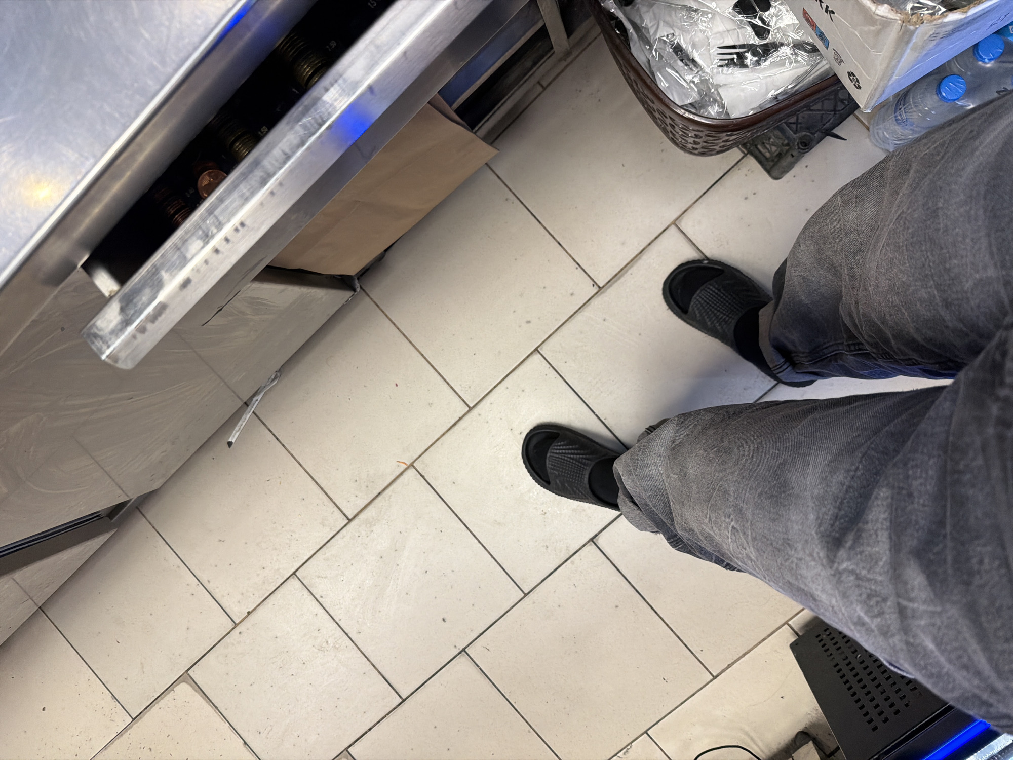 The image depicts a first-person perspective of a person standing on a tiled floor, likely indoors. The person is wearing dark grey jeans and black slides with socks. To the right, a basket containing plastic cutlery wrapped in plastic is visible, along with a box of bottled water. In the background, a metallic counter-top appliance is present, with what appears to be coins in a slot and a brown paper bag underneath. The overall environment suggests a retail or service area, possibly a shop or a food establishment. The location is specified as Plovdiv, Bulgaria.