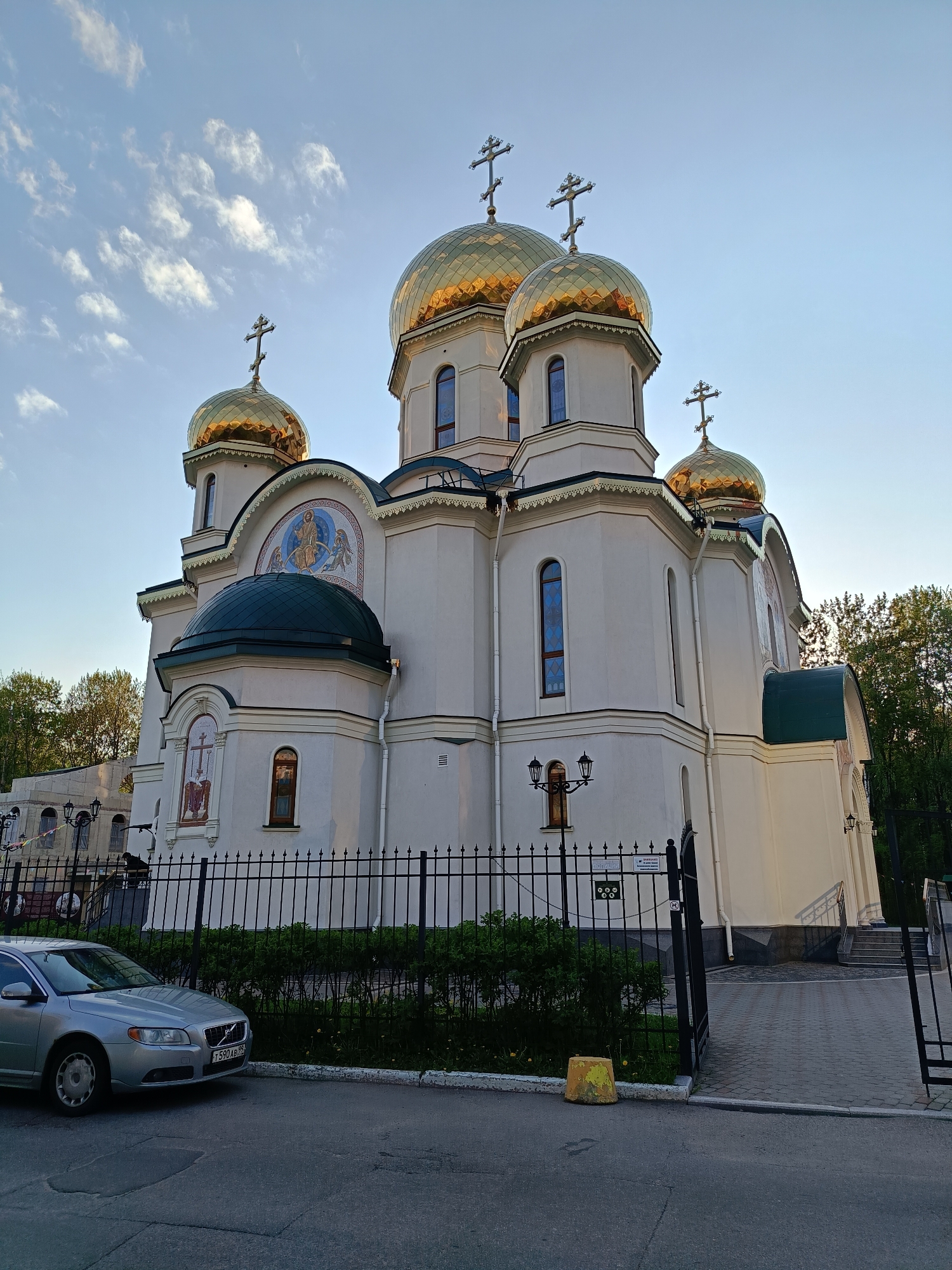 A ground-level, full, color photograph features a cream-colored Russian Orthodox church with multiple gilded onion domes under a blue sky with scattered clouds. The church is enclosed by a black metal fence with a gate slightly ajar. In front of the fence, on a paved street, is a parked silver Volvo car. The car's license plate is partially visible.
Behind the church, green trees are visible. The church's domes are topped with Orthodox crosses. The church building is detailed with arched windows and decorative elements. Some of the windows are narrow and rectangular. There is a smaller dark green dome on the side of the church.
The scene is taken during daylight hours, with sunlight illuminating the front of the church. The location is the Saint Petersburg Cathedral of the Icon of the Mother of God "Joy of All Who Sorrow" in Saint Petersburg, Russia.
