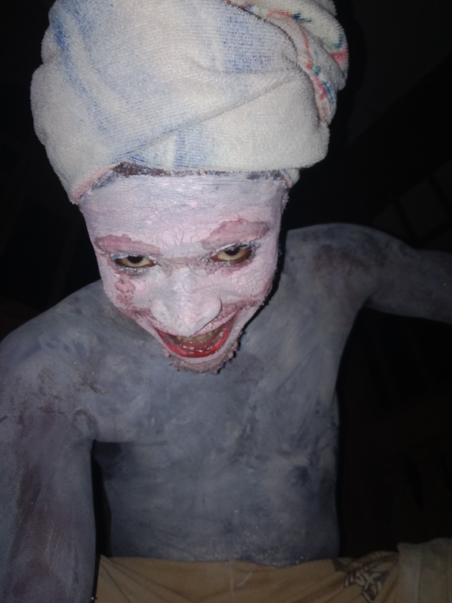 A person with gray and pink paint covering their face and body, and a white towel wrapped around their head, is depicted in a close-up, slightly high-angle shot. The person has a wide, unsettling smile that reveals their teeth, and their eyes are focused intently toward the viewer. The face paint is cracked, and there's a slight redness around the eyes and mouth. The background is dark and out of focus. The person is wearing light-colored pants. Location: Unknown, Unknown.