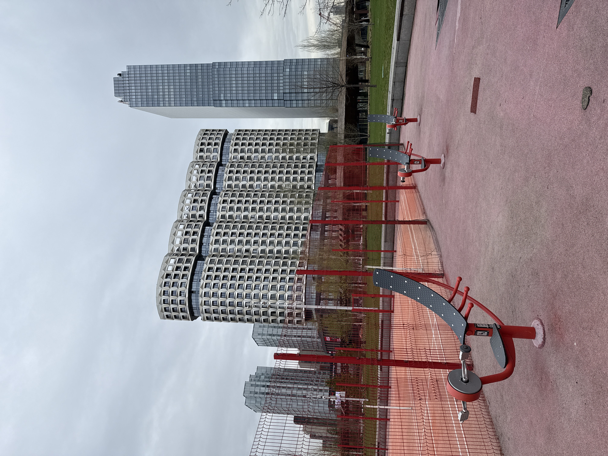 The image depicts an outdoor exercise area in Moscow, Russia, on an overcast day.
In the background, two modern skyscrapers dominate the skyline. One has a conventional rectangular design, while the other is characterized by a more unusual, undulating structure featuring repeating patterns of windows.
In the foreground, several pieces of red and black outdoor exercise equipment are positioned on a red rubberized surface, partially obscured by a red metal fence.