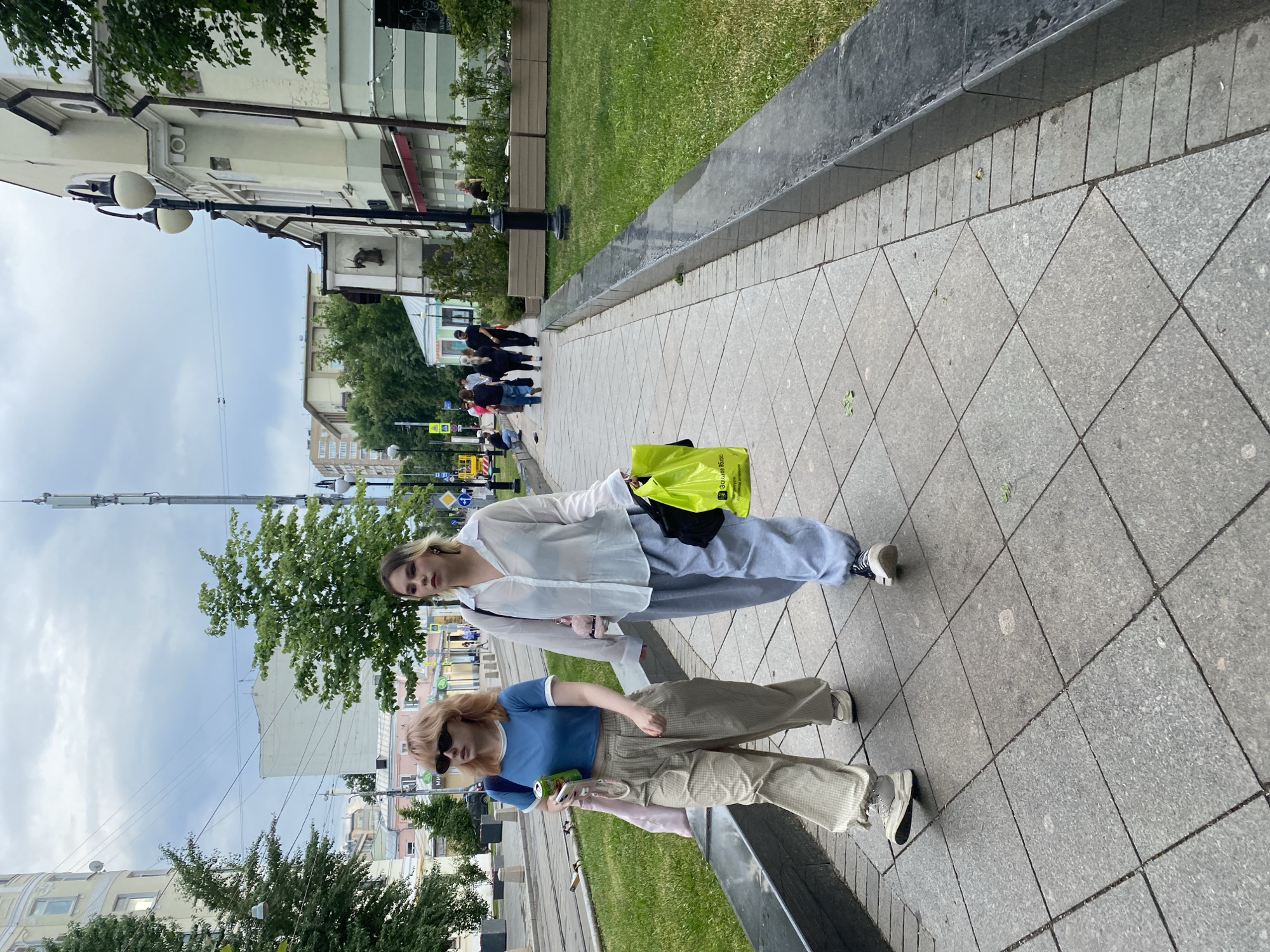 Two young women are walking side-by-side on a pedestrian sidewalk in Moscow, Russia. The woman on the left has blonde hair and is wearing a blue crop top, beige checkered pants, and white sneakers. She is holding a green can and a pink bag. The woman on the right has blonde hair with darker roots and is wearing a white loose-fitting shirt, gray sweatpants, and black and white sneakers. She is carrying a bright yellow shopping bag. They are walking away from the camera. The sidewalk is paved with gray square tiles. To the left of the sidewalk is a grassy area with trees and a street with tram tracks. To the right of the sidewalk is a building with a dark facade and several windows, some of which are covered with awnings. The sky is overcast. In the background, other pedestrians can be seen walking, as well as buildings and a cell tower. The scene appears to be a street in a city with commercial and residential buildings.