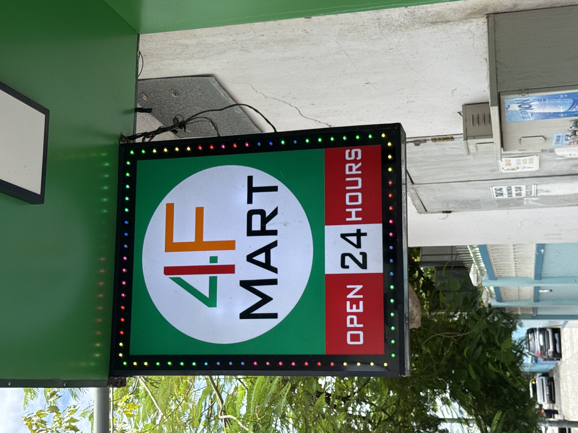 An illuminated sign for "4F MART" is prominently displayed, featuring the store's name in large, bold letters against a green background, with "4.F" in red and orange within a white circle. Below this, the words "OPEN 24 HOURS" are visible in white text on a red and white segmented background. The sign is outlined by colorful LED lights. The sign is attached to a green awning extending from a building with a textured, light-colored wall. The background includes lush green foliage and a glimpse of a street scene with cars. The scene appears to be an outdoor commercial area in Nha Trang, Vietnam, characterized by the architecture and vegetation.
